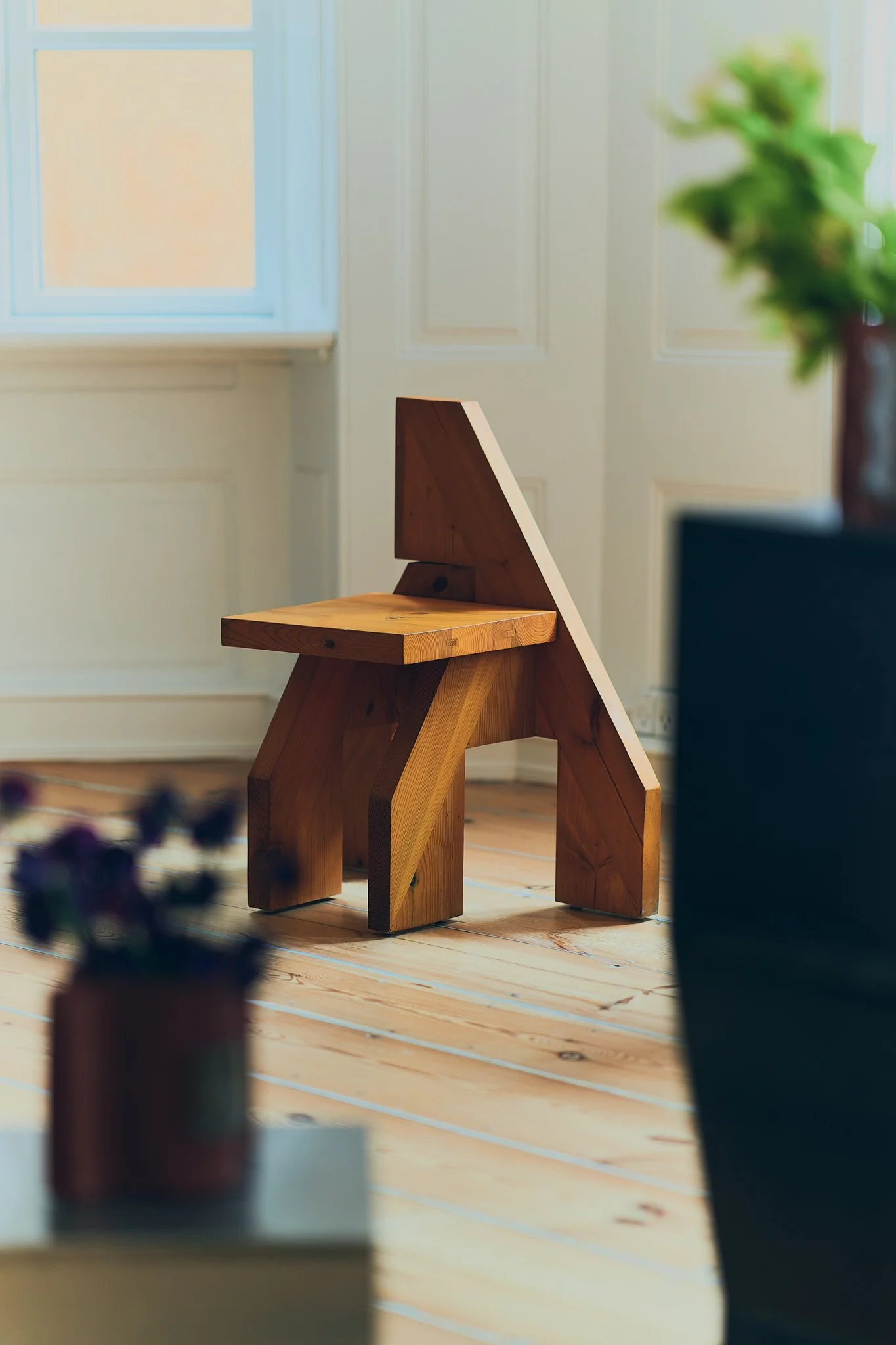 Wooden chair with geometric design in a room with wooden floors, near a window, and blurred foreground plant.