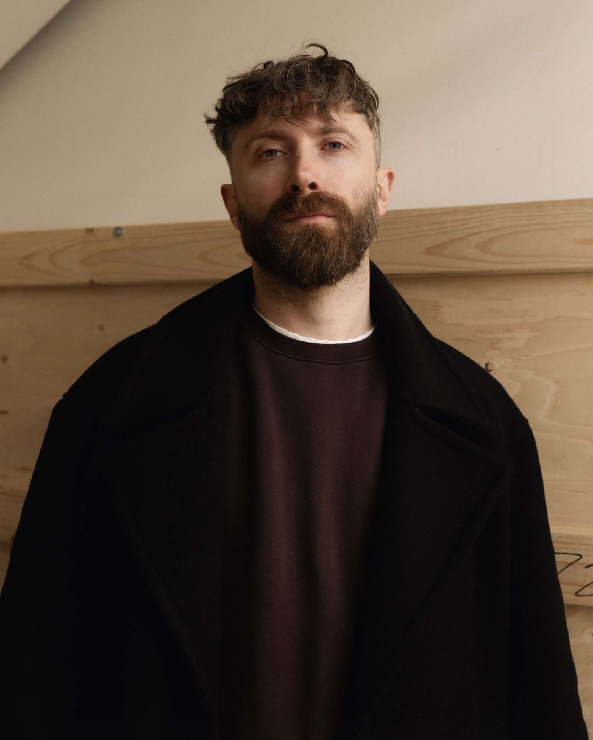 Man with a beard wearing a black coat and standing against a wooden background.