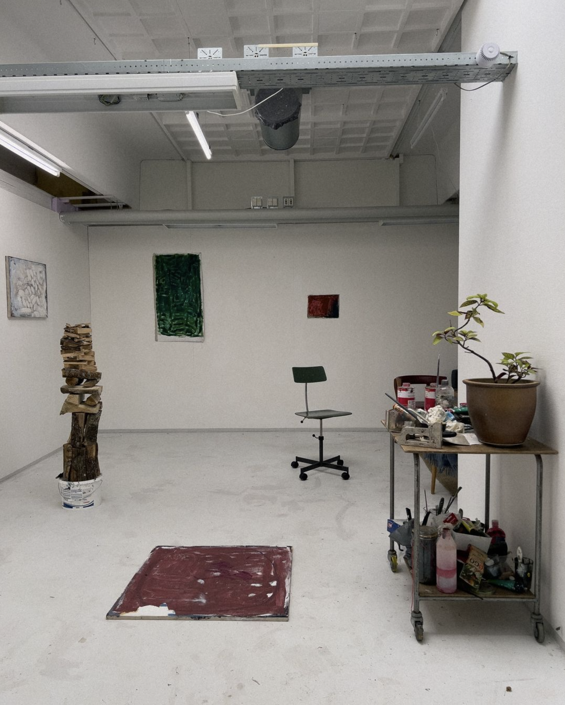 Minimalist art studio with a chair, artworks on the floor and walls, a sculpture of stacked wood, and a cart with art supplies and a potted plant.