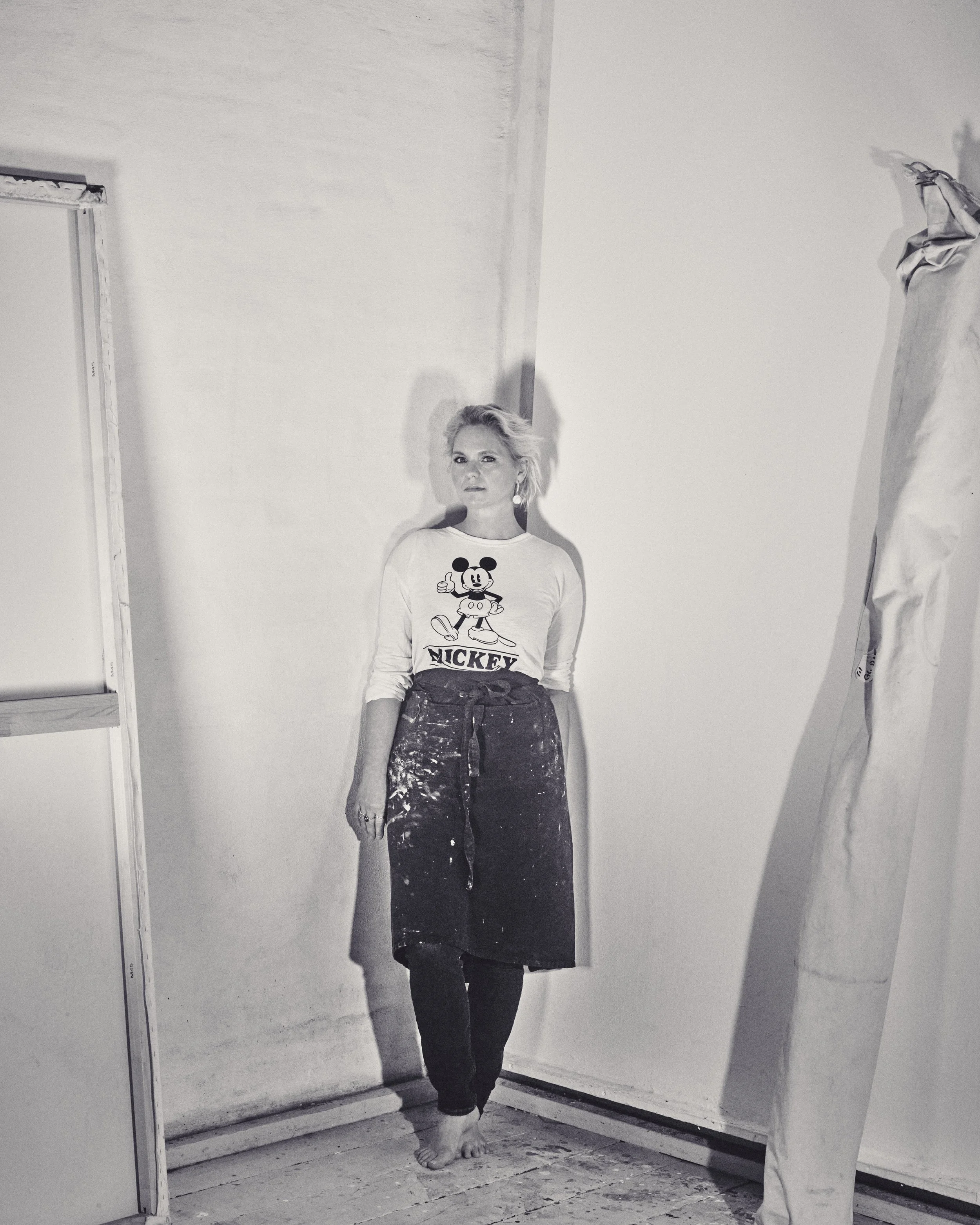 Black and white photo of a woman leaning against a wall, wearing a Mickey Mouse shirt and apron, with paint stains on the floor and an unrolled canvas or paper nearby.
