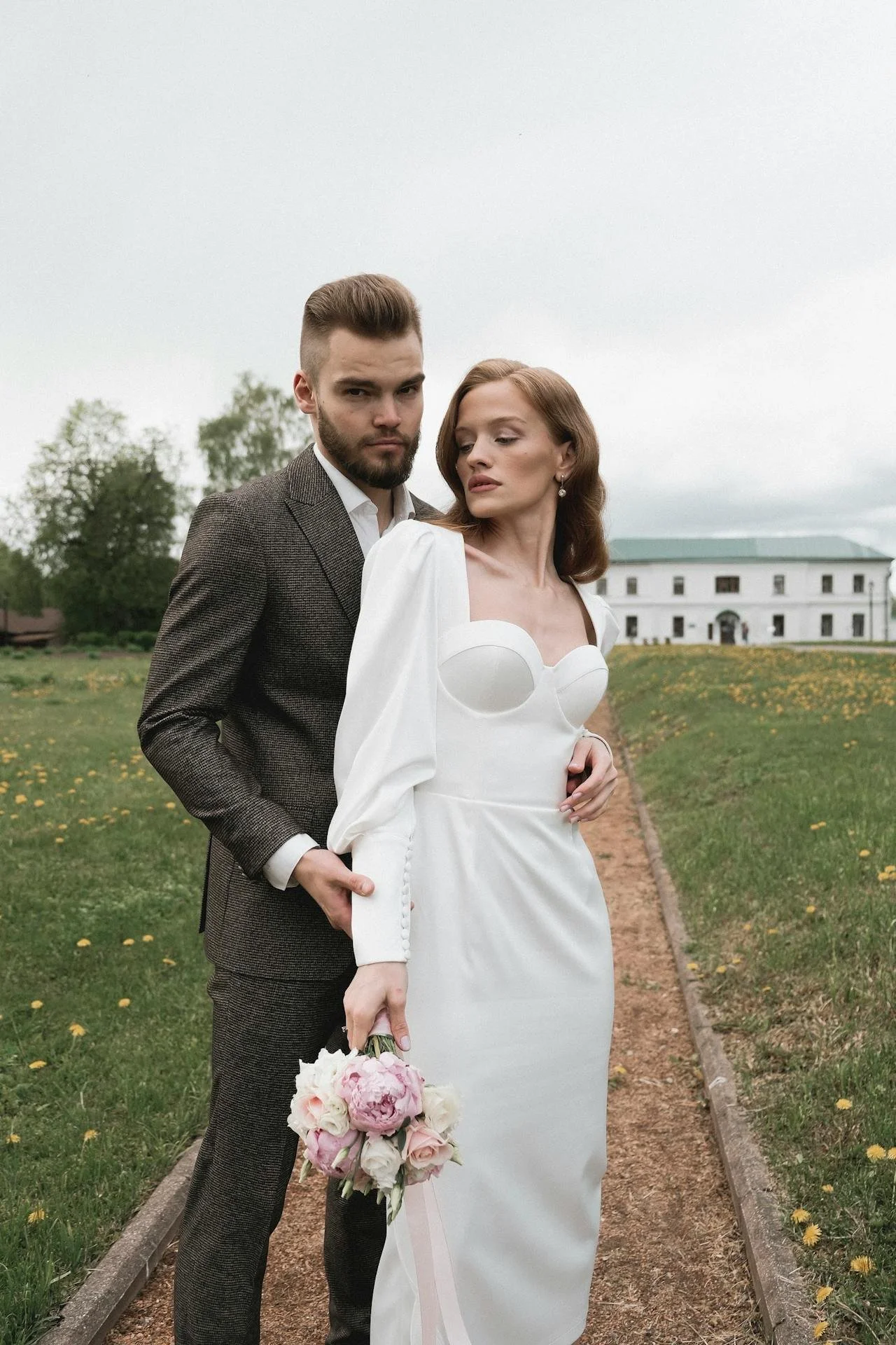A couple in formal attire standing outdoors on a pathway. The woman is in a white dress holding a bouquet of flowers, and the man is in a dark suit. They appear to be posing for a formal photo, with greenery and a building in the background.