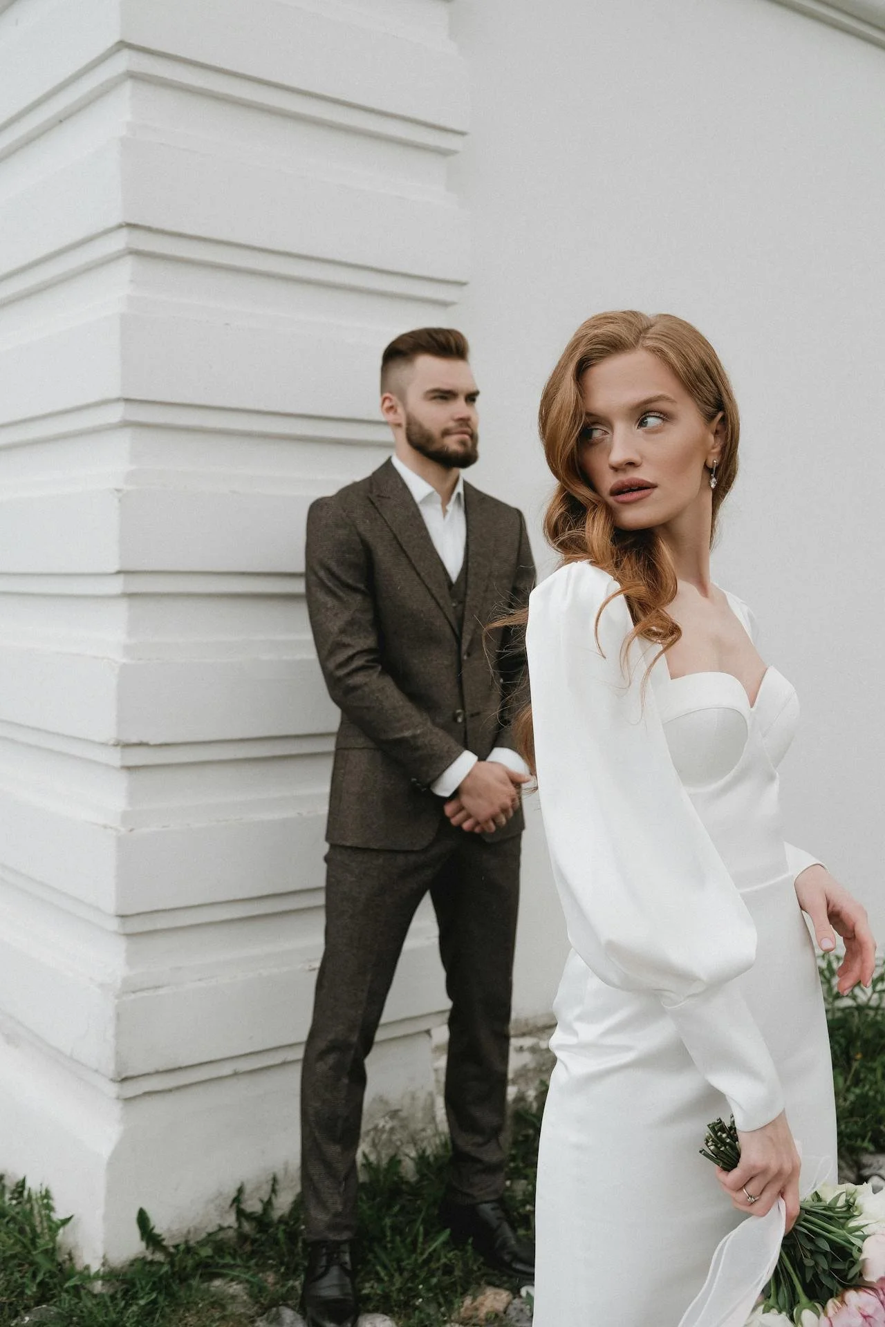 Wedding Website Design, Digital Wedding Stationery, Web Design and Branding for Wedding Professionals | Avelã White. Bride in a white wedding dress holding flowers, standing in front of a groom in a brown suit against a white wall.