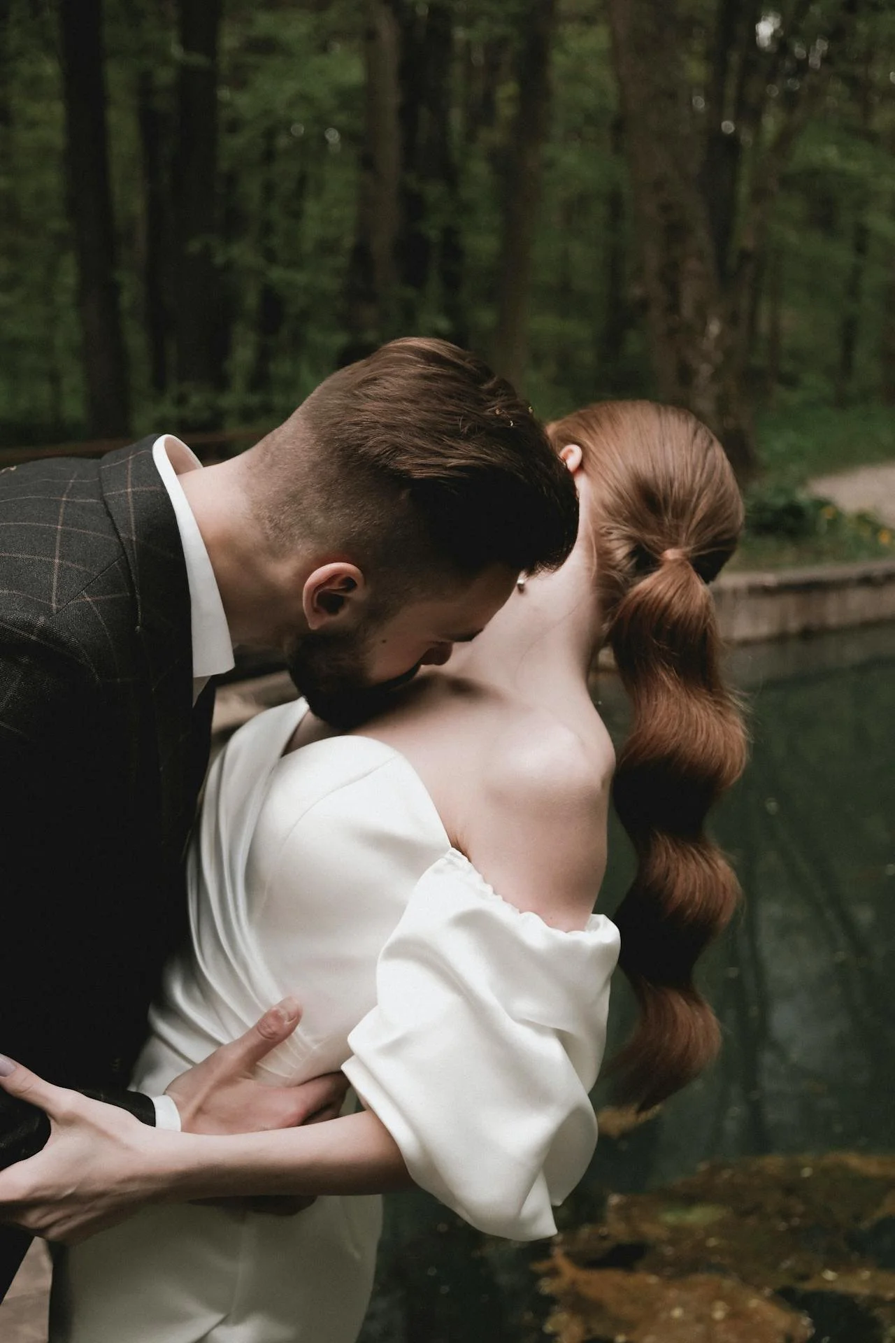 Wedding Website Design, Digital Wedding Stationery, Web Design and Branding for Wedding Professionals | Avelã White. Man in dark suit kissing a woman in a white dress near a pond in a forest setting.