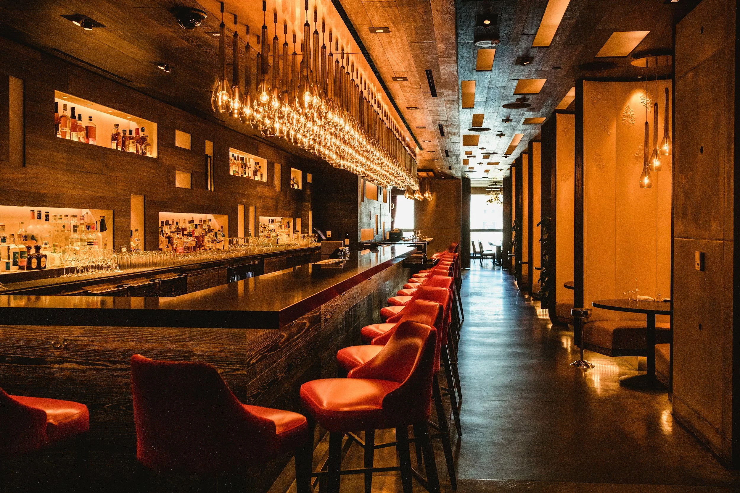 A cozy bar with a long, dark wooden counter lined with red leather chairs, illuminated by warm hanging pendant lights, with a well-stocked backlit liquor shelf, and a seating area near windows in the background.