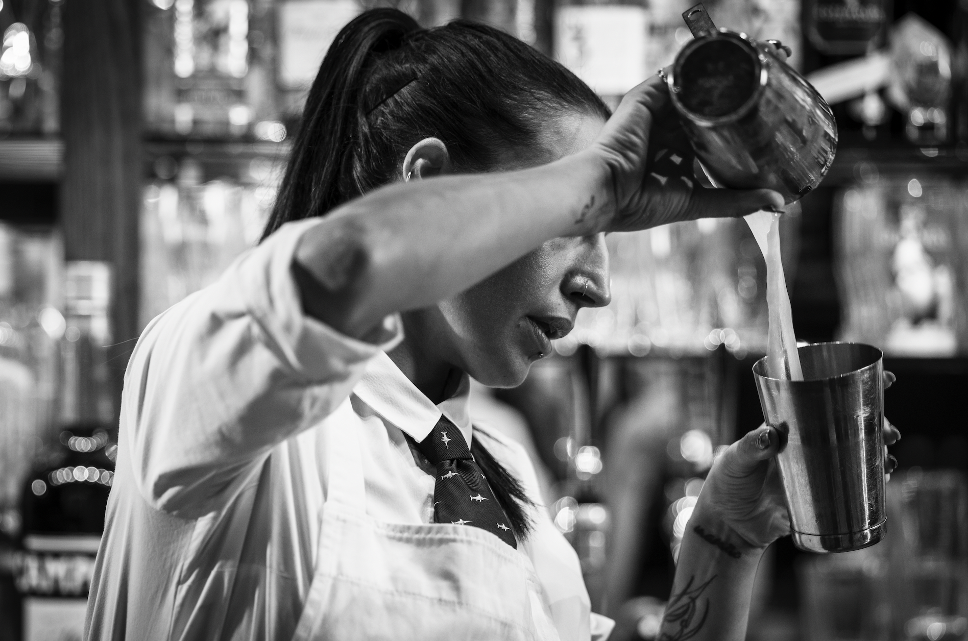 A woman with a ponytail pouring a liquid from a small container into a metal cup in a busy kitchen or bar setting.