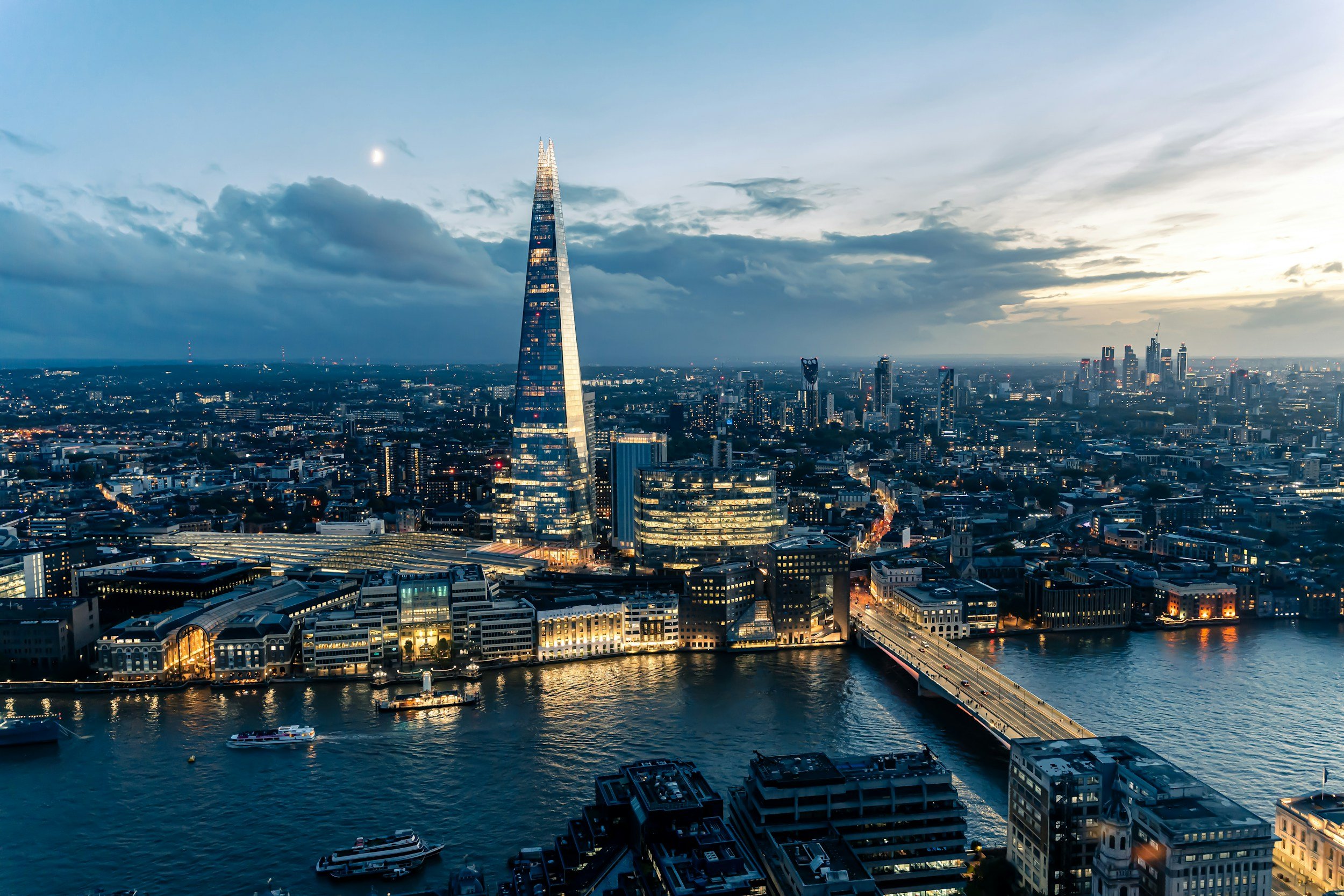 Skyline of London at dusk with The Shard prominent, Thames River in foreground, city lights beginning to illuminate, and cloudy sky overhead.