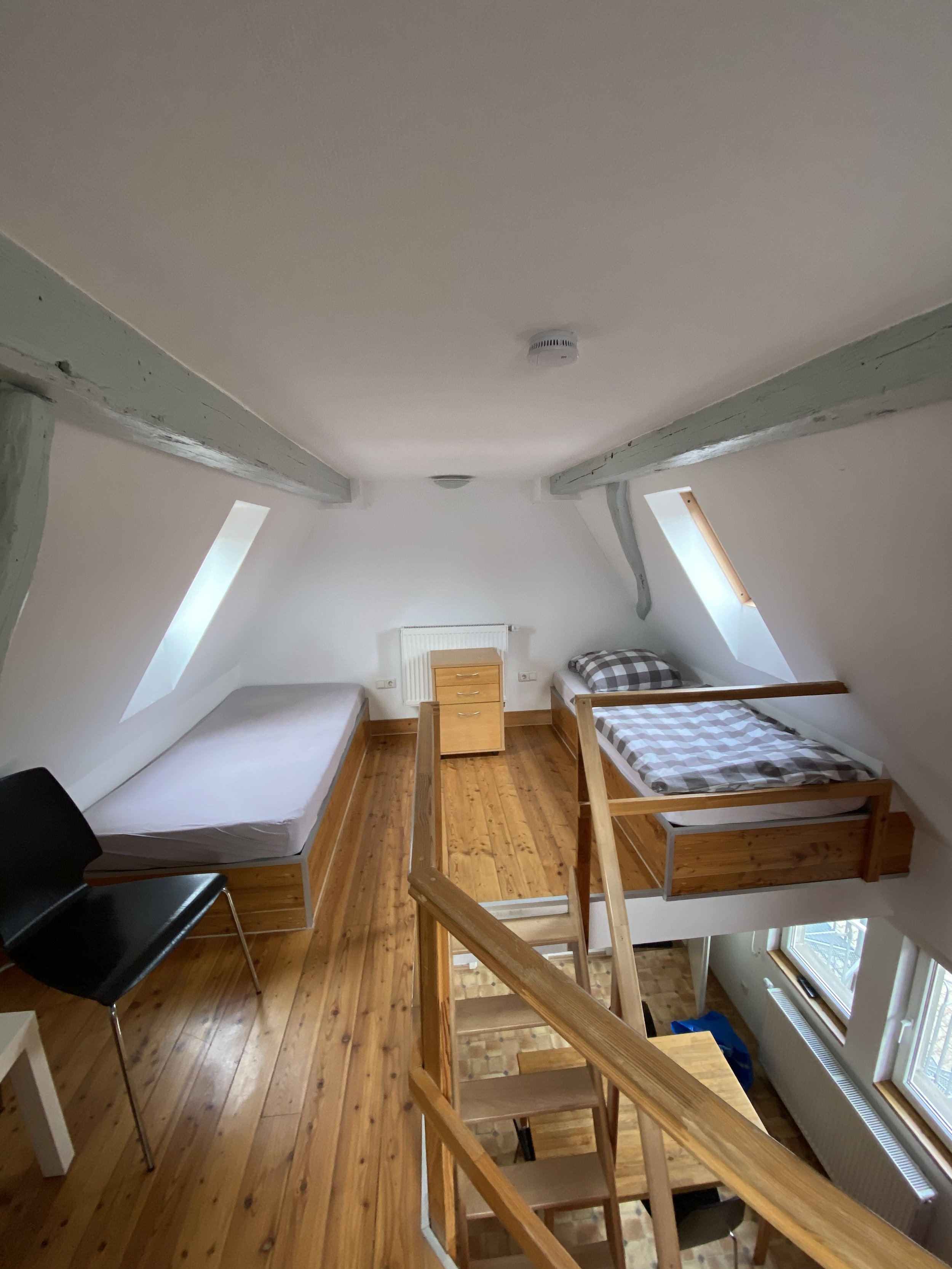 Ein loftartiges Schlafzimmer mit zwei Betten, einer Treppe, Holzfußboden, weißem Decke, Fenster und Möbeln in hellen Holzfarben.