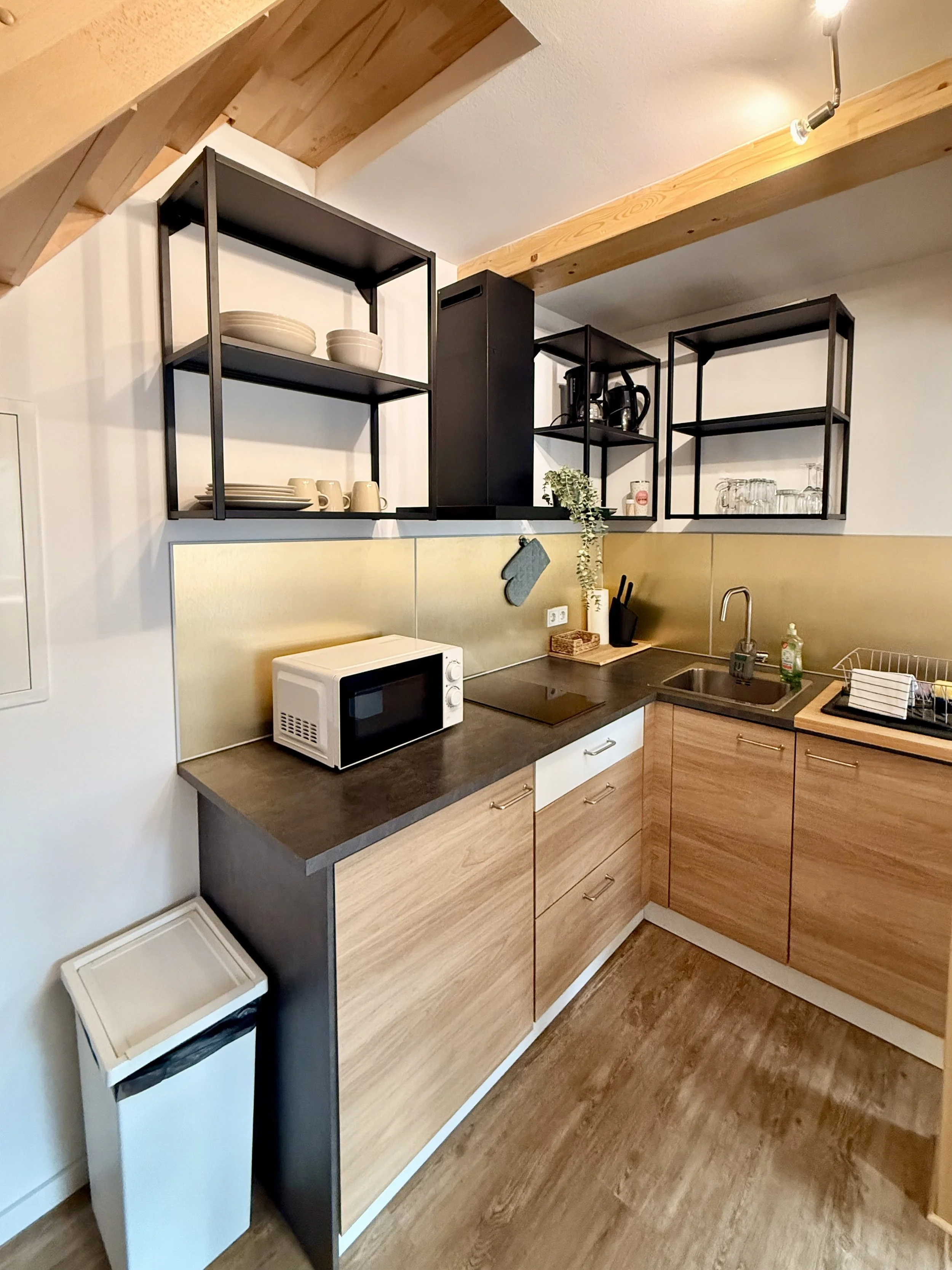 Küche mit Holzunterschränken, schwarzen offenen Regalen, weißen Geschirr und einem Mikrowellenherd auf der Arbeitsplatte.