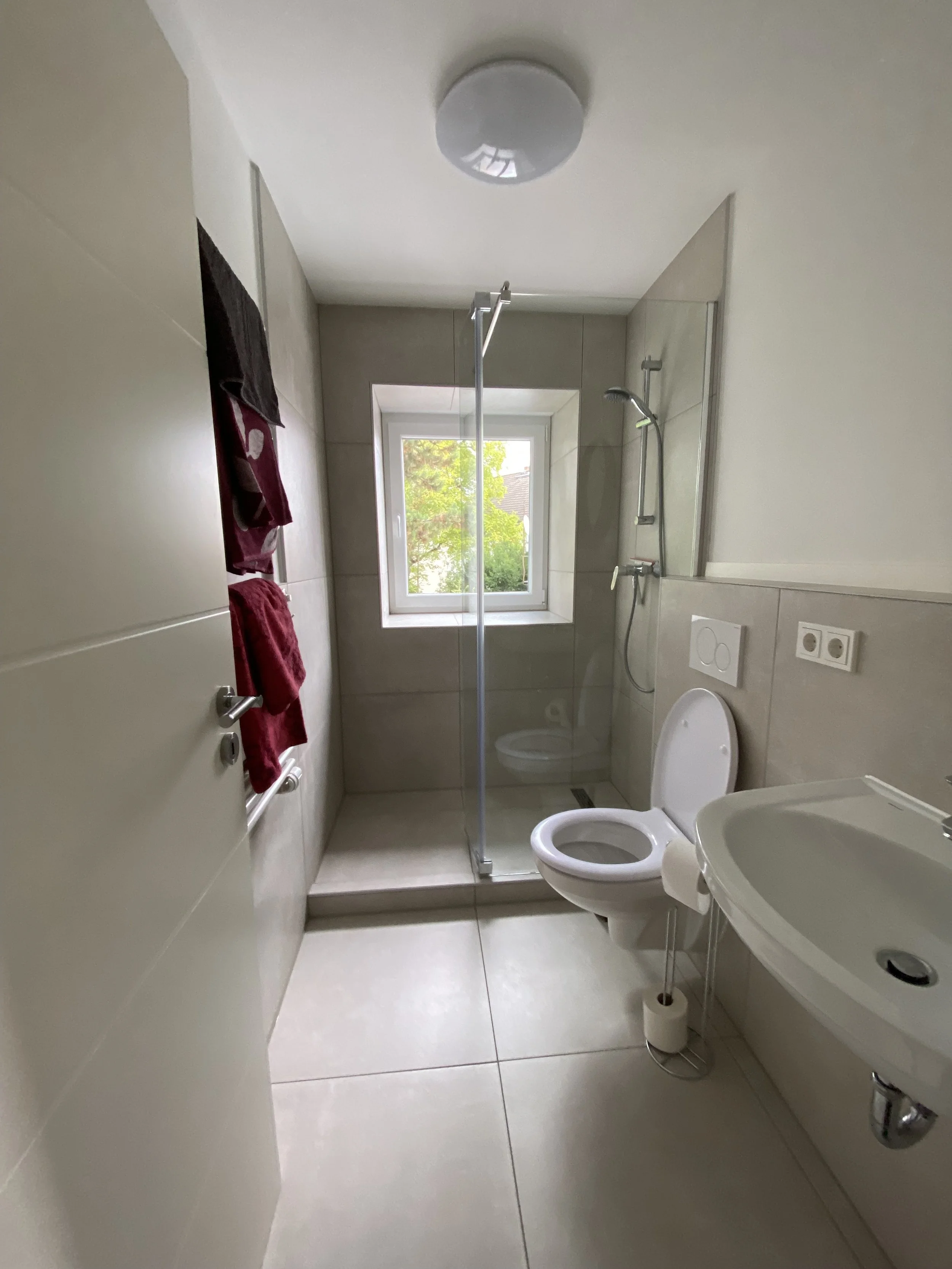 Ein Badezimmer mit Dusche, WC und Waschbecken, Fenster mit Blick nach draußen, Handtücher an einem Handtuchhalter, weiße Fliesen an den Wänden und Böden.