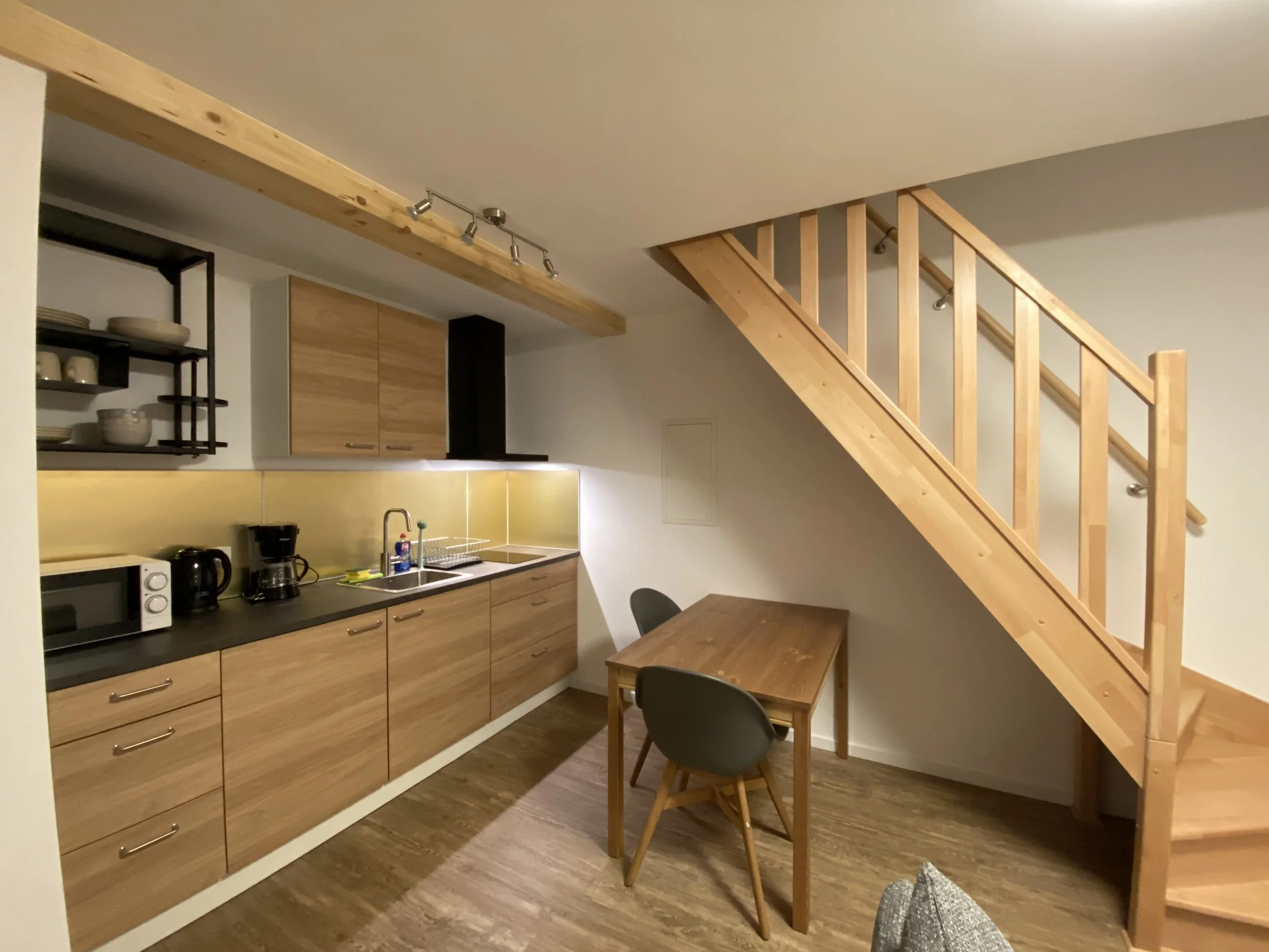Küche und Esstisch unter einer Holztreppe, helle Holzmöbel, dunkle Stühle, Küche mit Geschirr, Mikrowelle, Kaffeemaschine, Herd, Spüle, offene Regale