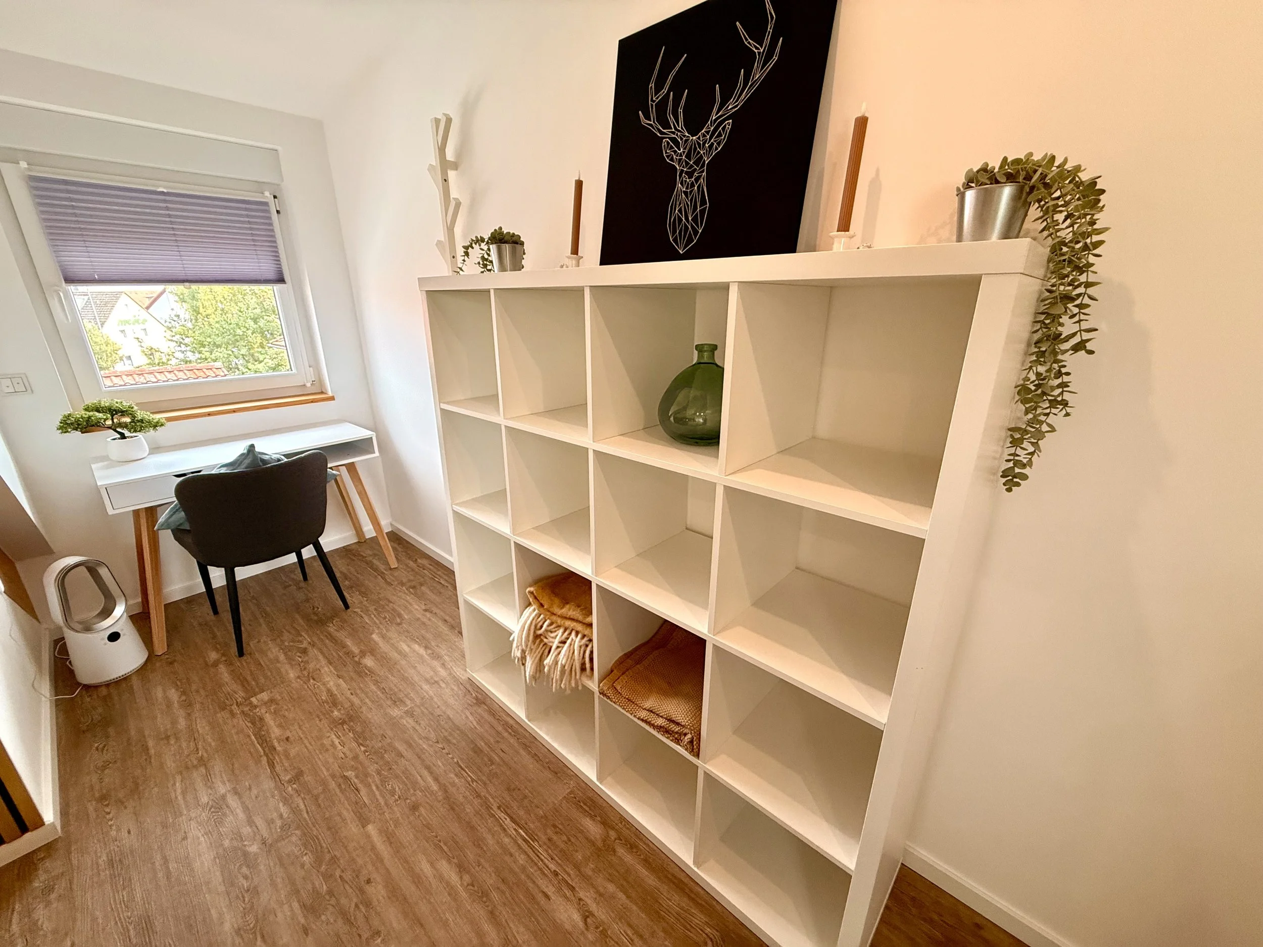 Ein kleines Arbeitszimmer mit einem weißen Schreibtisch neben einem Fenster, einem schwarzen Stuhl, einem weißen Wandkühlschrank, weißen Regalen mit Dekorationsobjekten, Pflanzen und einem Bild mit Hirschkopf auf dem Regal.