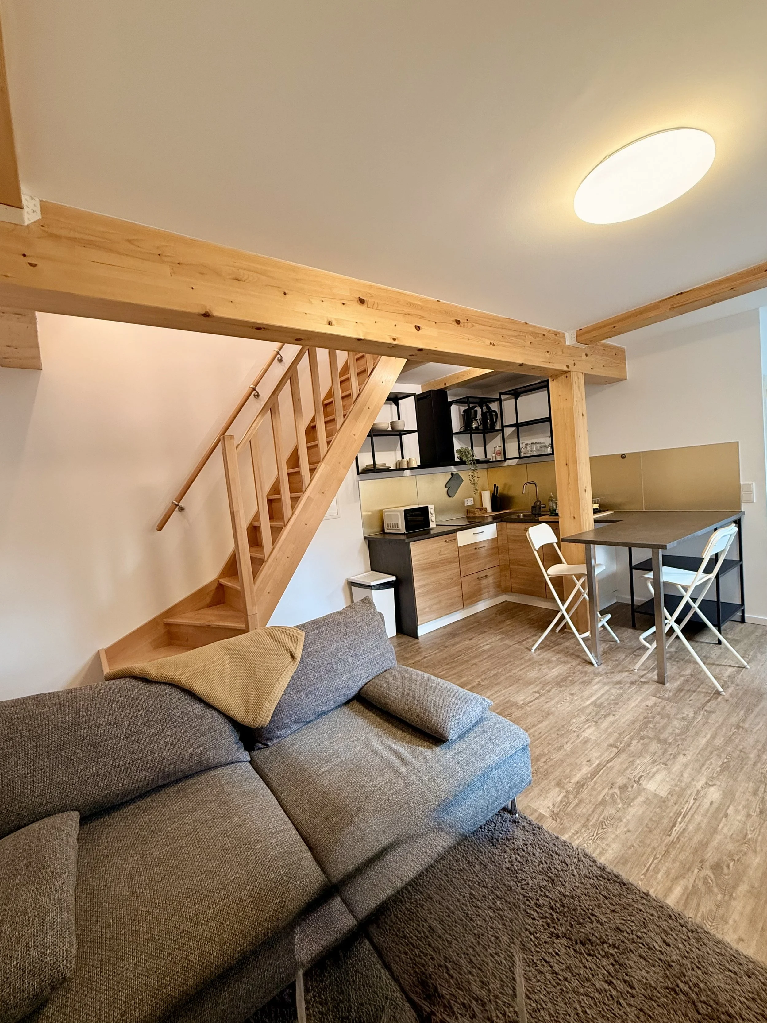 Ein offener Wohnbereich mit einer Couch, einer Küchenzeile, einem Esstisch und Treppe zu einer oberen Etage, alle im modernen Stil mit Holz- und Neutralfarben.