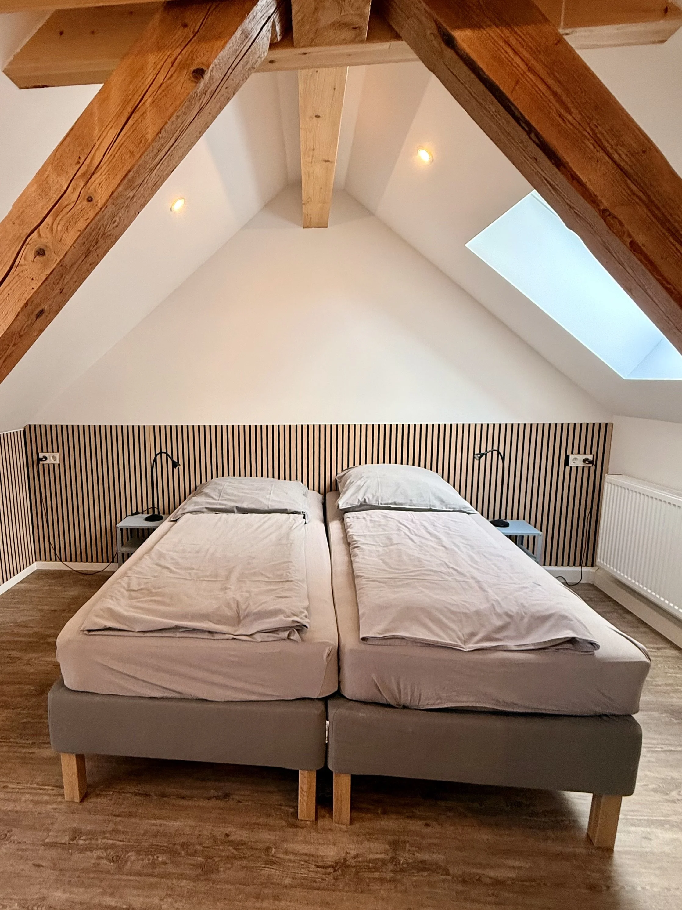 Ein Schlafzimmer mit zwei Betten, Holzfußboden, schräg verlaufende Decke mit Dachfenster, Holz-Akzentwand hinter den Betten, Leselampen neben den Betten, seitliche Nachttische.