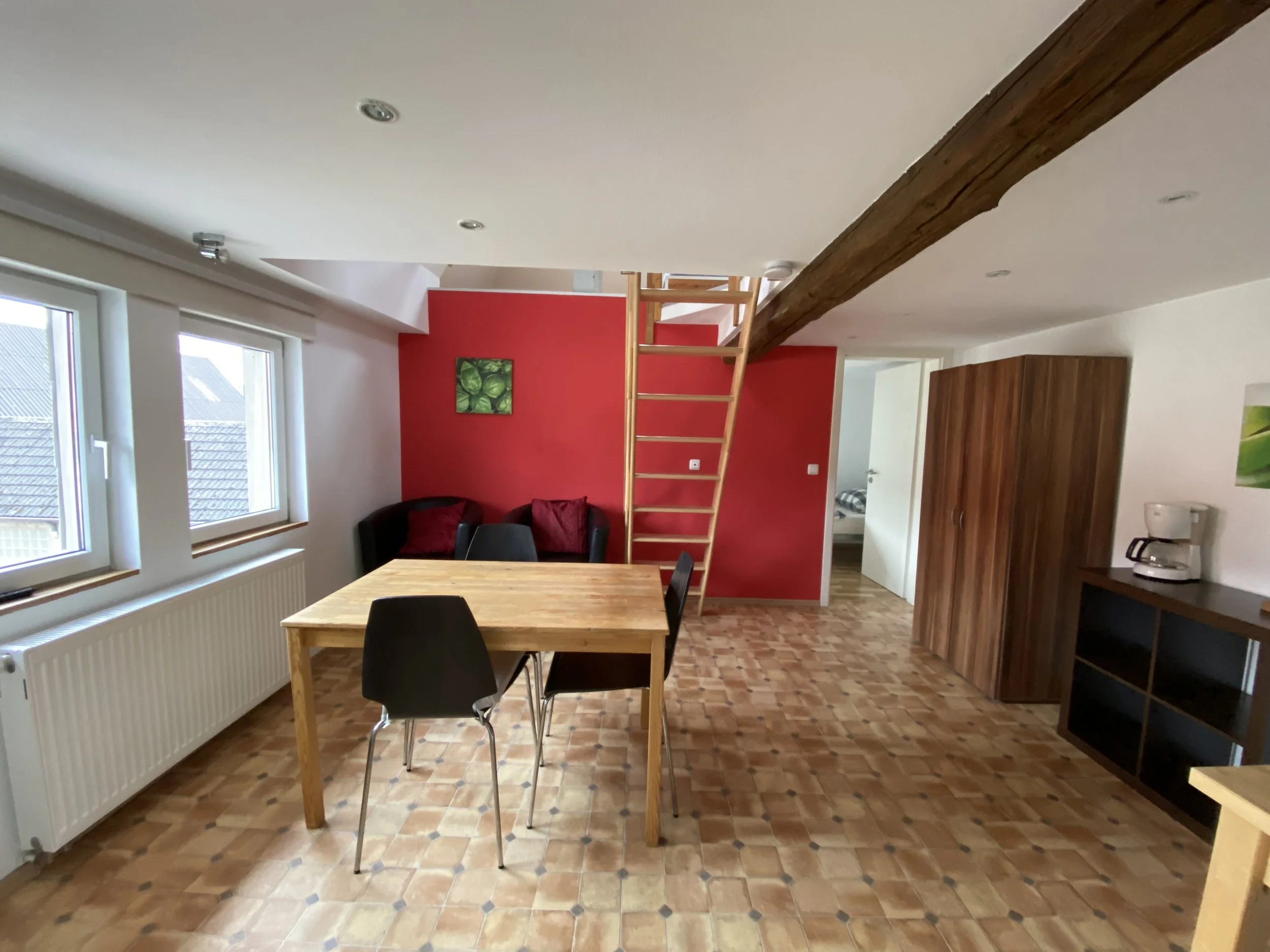 Ein Wohnzimmer mit roter Wand, Holzfußboden, Esstisch mit vier schwarzen Stühlen, dunkelgrauer Sessel mit roten Kissen, offenes Treppenhaus mit Holztreppe, moderner Einrichtungsstil.