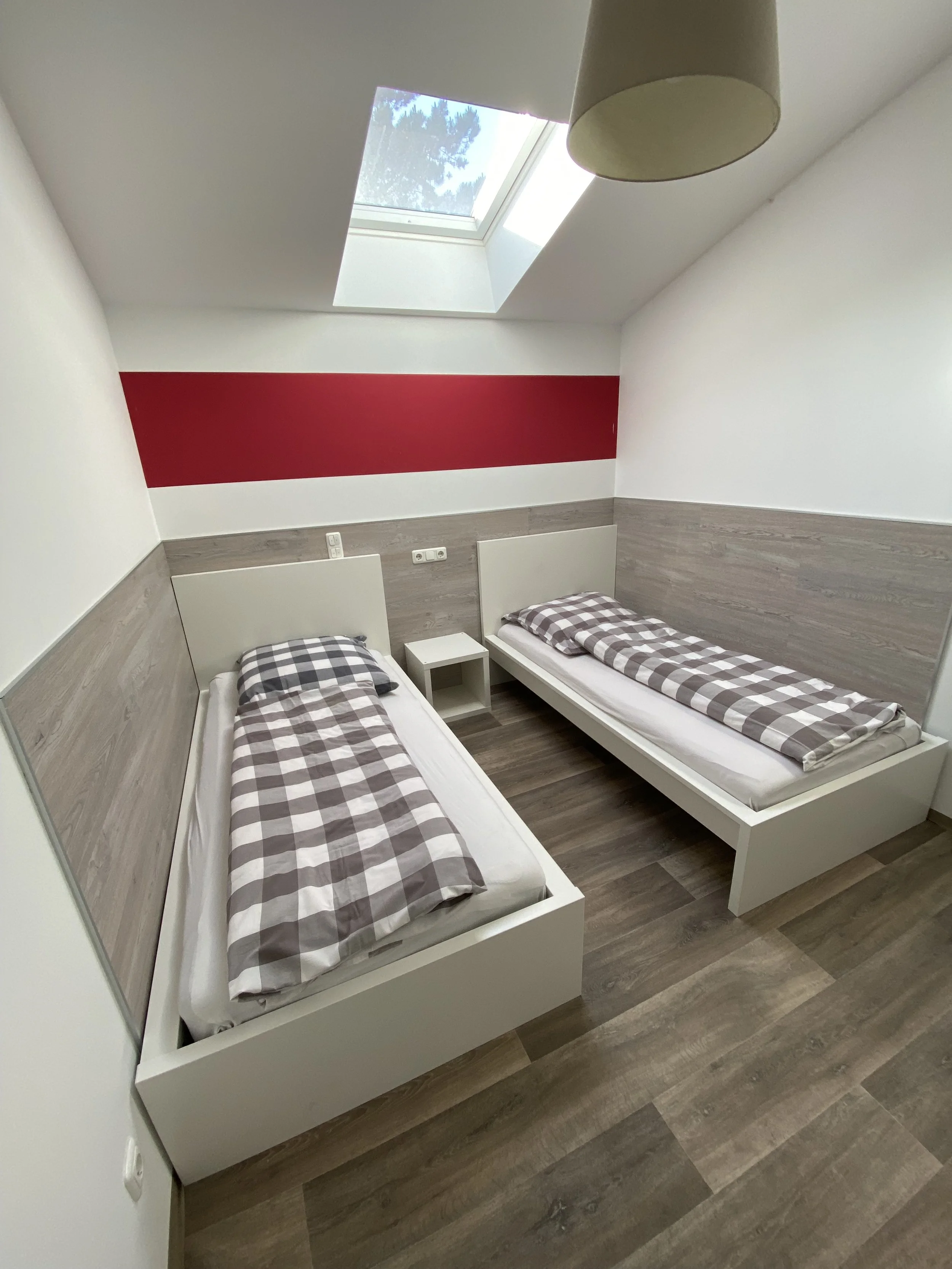 Ein Zimmer mit zwei Einzelbetten, einer kleinen Nachttisch und einem Dachfenster mit Sicht auf den Himmel und Bäume.