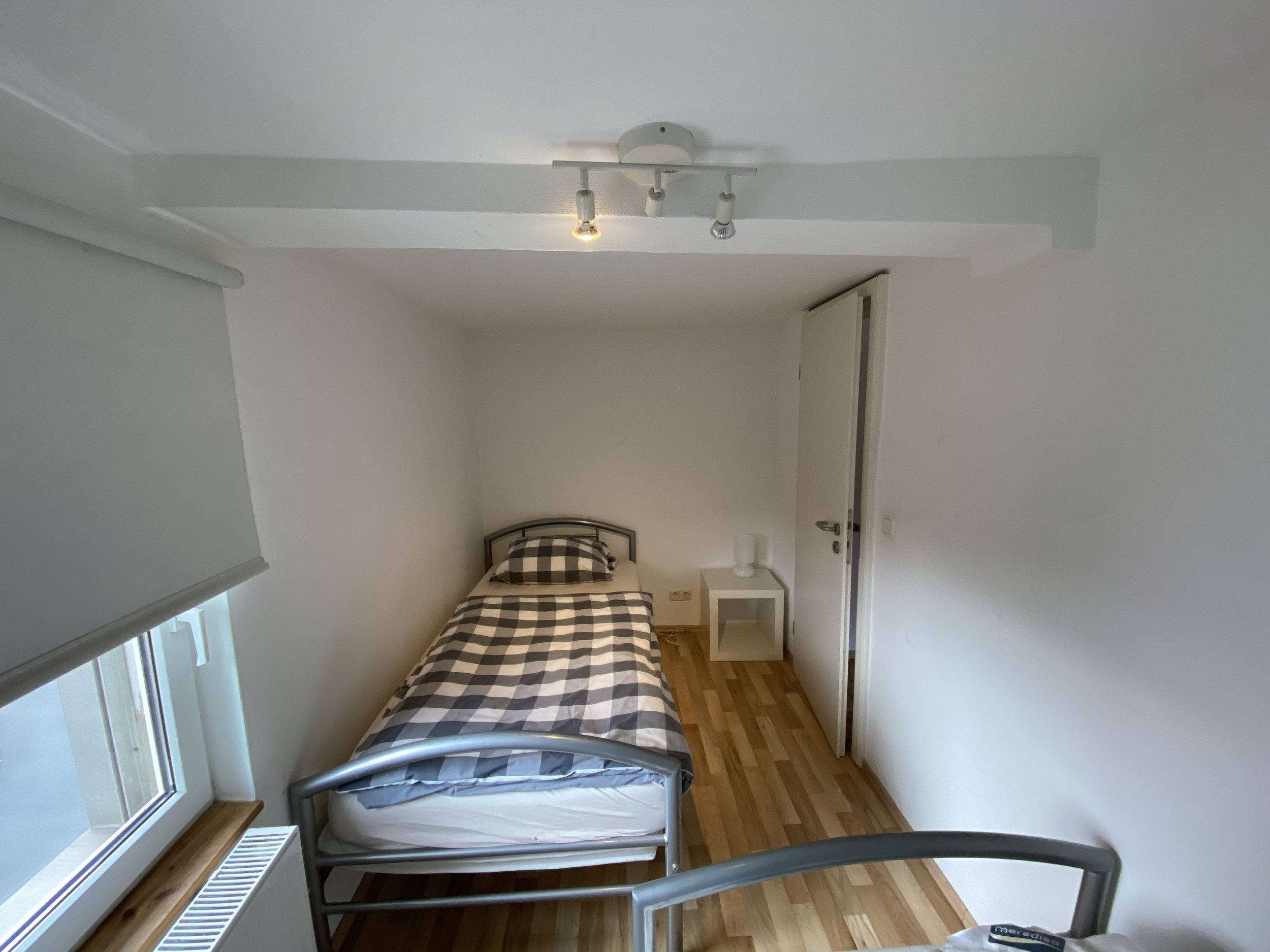 Kleine, schlichte Schlafzimmer mit Einzelbett, Nachttisch und Lampe, Holzboden, Stromkabel, Fenster mit Verdunkelungsrollo, weiße Wände, Deckenleuchte.