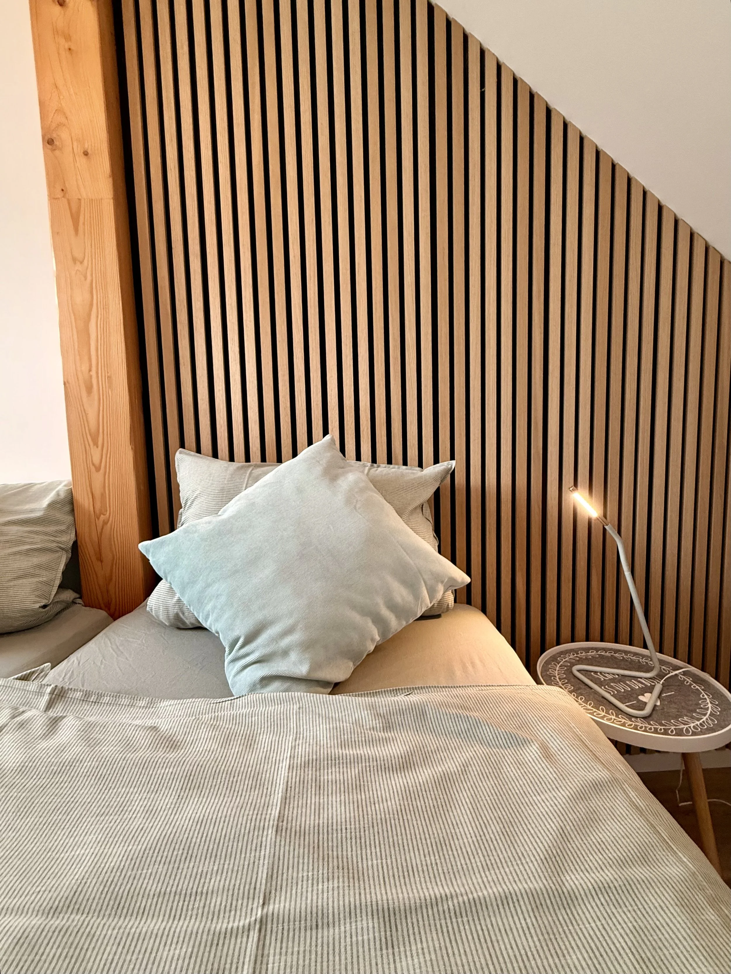 Gemütliches Schlafzimmer mit gestreiftem Bettzeug, mehreren Kissen und einem Holz-Lattenkopfteil. Neben dem Bett steht ein indirekt beleuchteter Nachttisch.