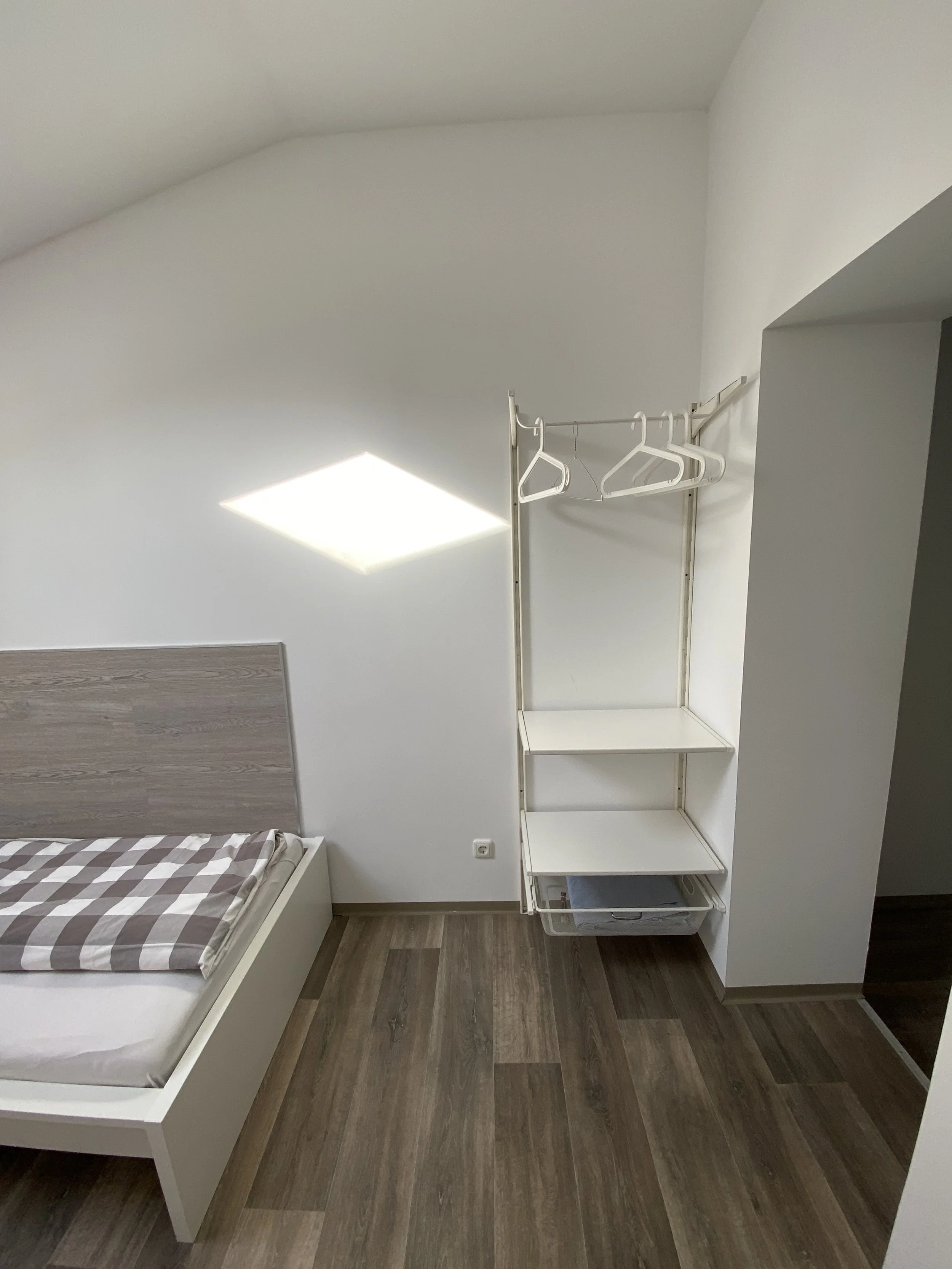 Ein Schlafzimmer mit einem weißen Bett, einem Holzfußboden, einer weißen Wand und einem offenen weißen Kleiderschrank mit Kleiderbügeln und Ablageflächen.