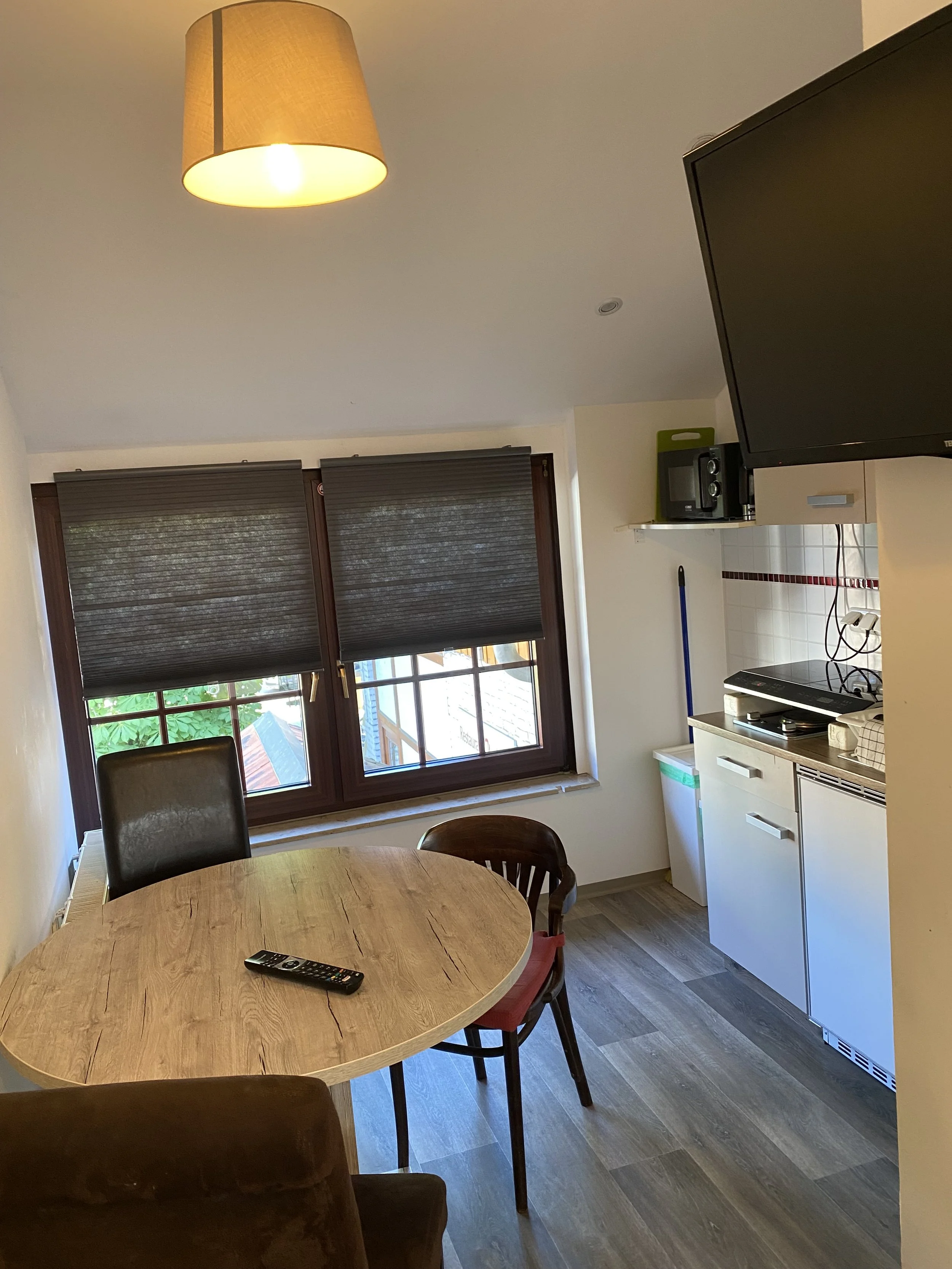 Ein Esszimmer mit einem runden Holztisch, mehreren Stühlen, einem Fenster mit halb gezogenen Jalousien, einer Deckenlampe, einer kleinen Küche mit Kochfeld, Mikrowelle, Geschirr und einem Becher, in einem hellen Raum.