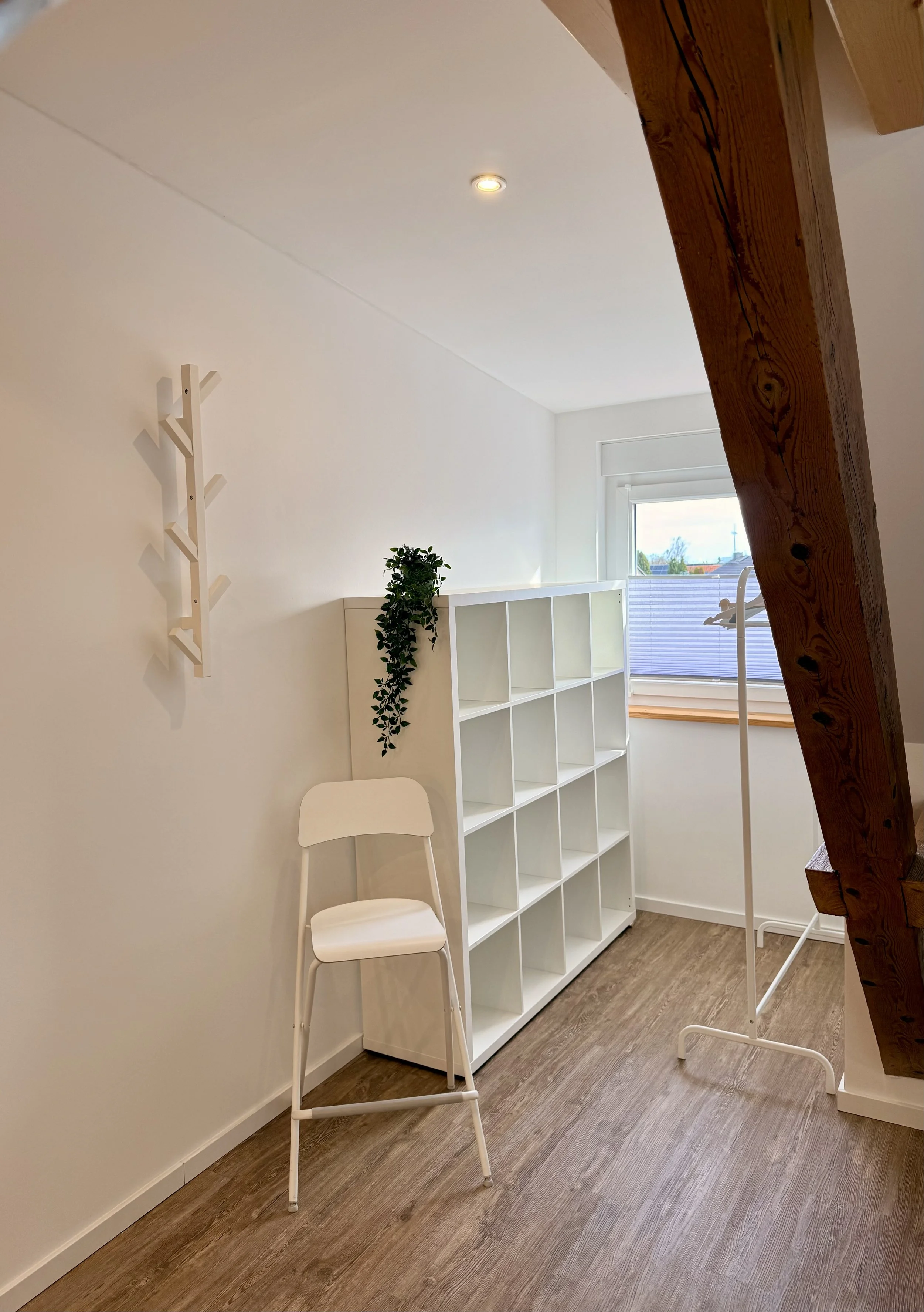 Ein weißes Regal neben einem weißen Stuhl, an der Wand eine hölzerne Wandhaken, vor einem Fenster hängt ein weißer Kleiderständer, im Raum Holzboden, eine Holzwandstütze im rechten Bild, natürliche Lichteinfälle aus dem Fenster.