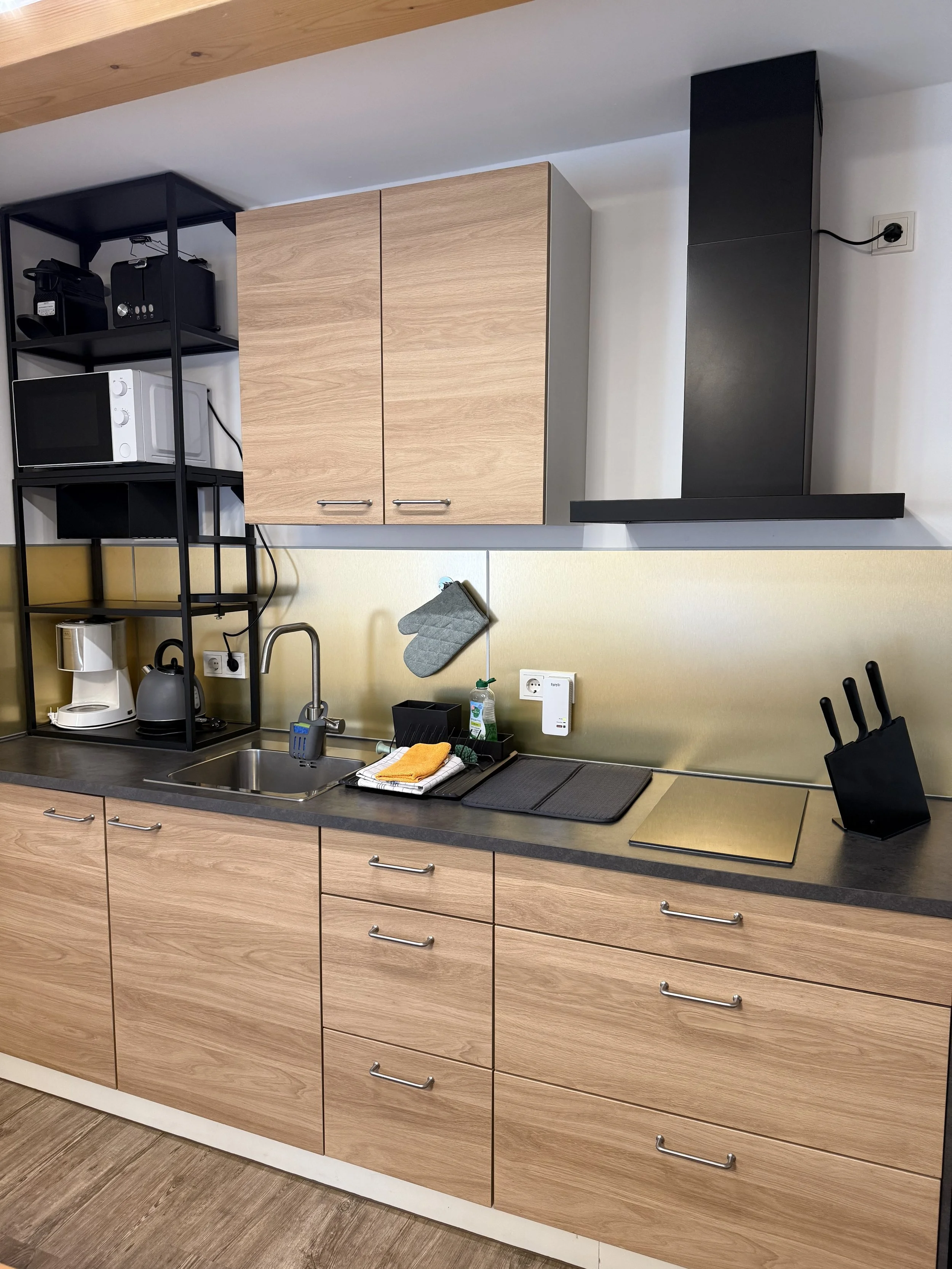 Küche mit Holzschränken, schwarzen Regalen, Mikrowelle, Wasserkocher, Kaffeemaschine, Spüle, Küchenutensilien, Herd und Messerblock.