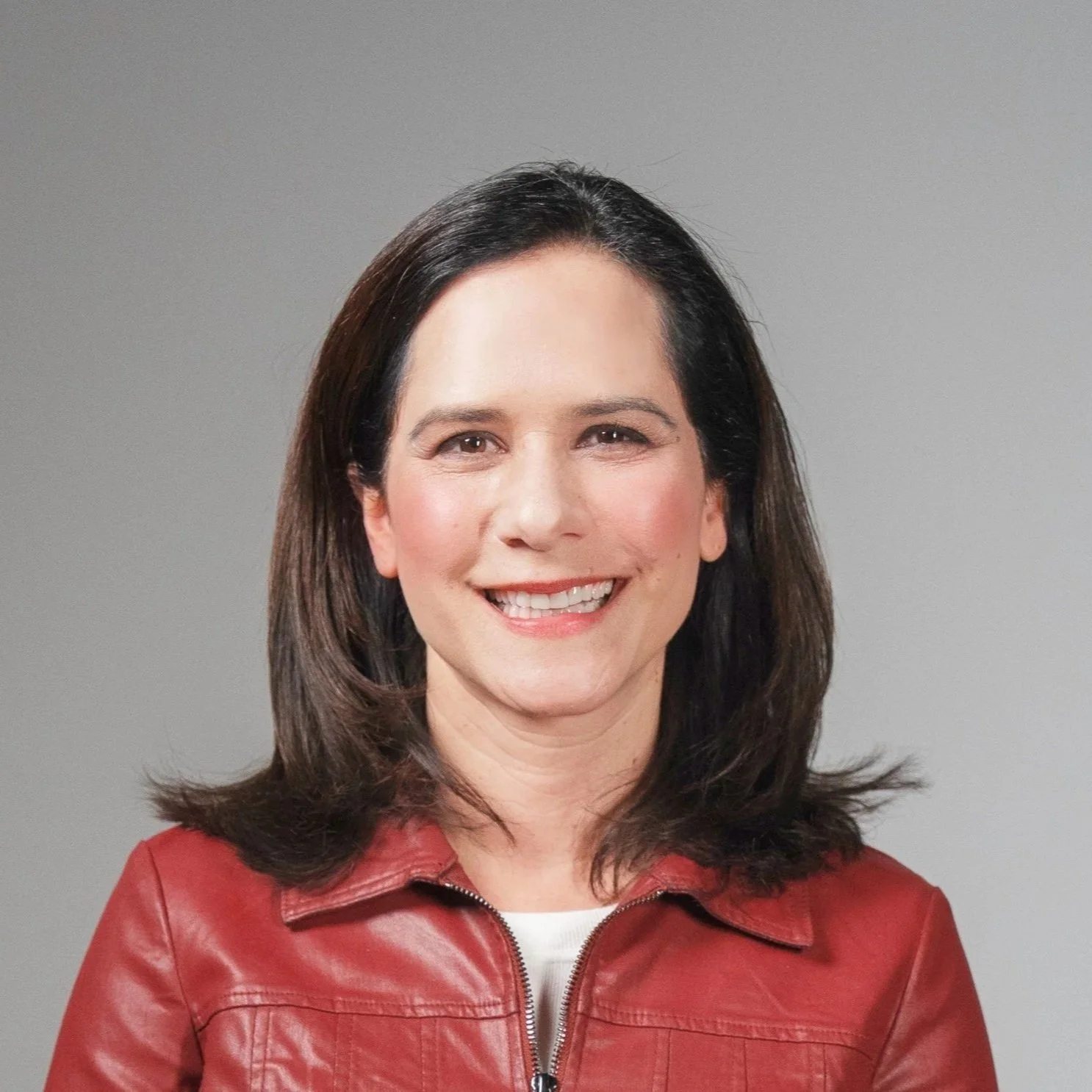 Portrait of a woman with dark brown hair, smiling, wearing a red leather jacket and a white top, against a plain gray background.