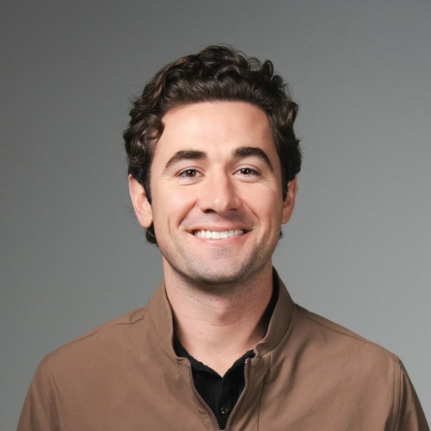 Headshot of a smiling man with dark, curly hair wearing a brown jacket over a black shirt, against a gray background.