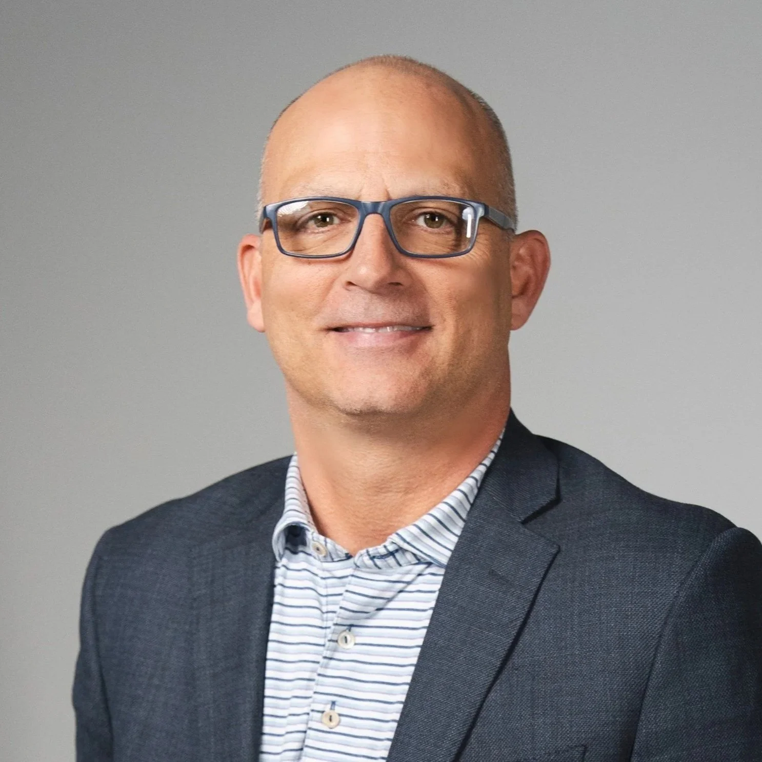 Professional headshot of a smiling middle-aged man with glasses, wearing a dark blazer over a striped shirt, against a gray background.