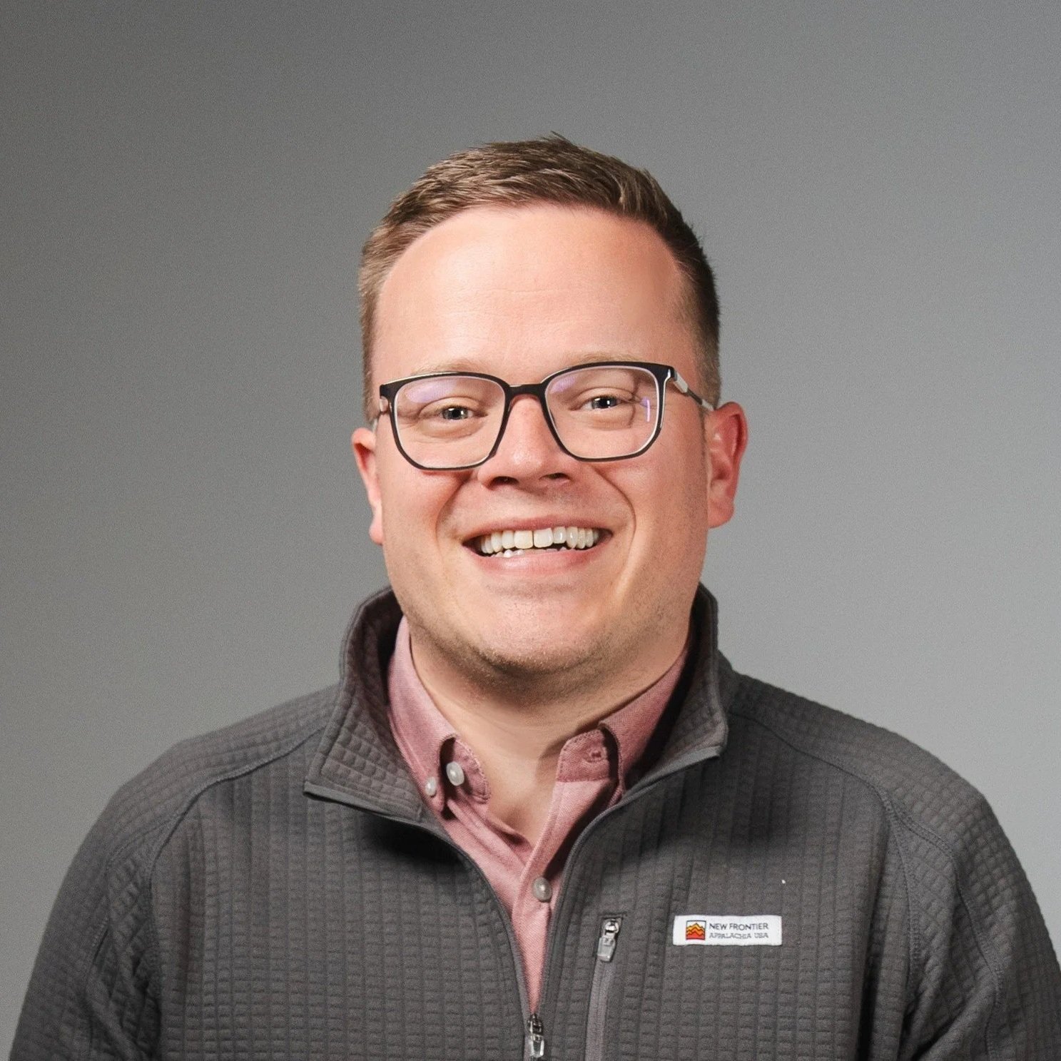 A smiling man with glasses wearing a gray zip-up jacket and a maroon collared shirt, posing against a plain gray background.
