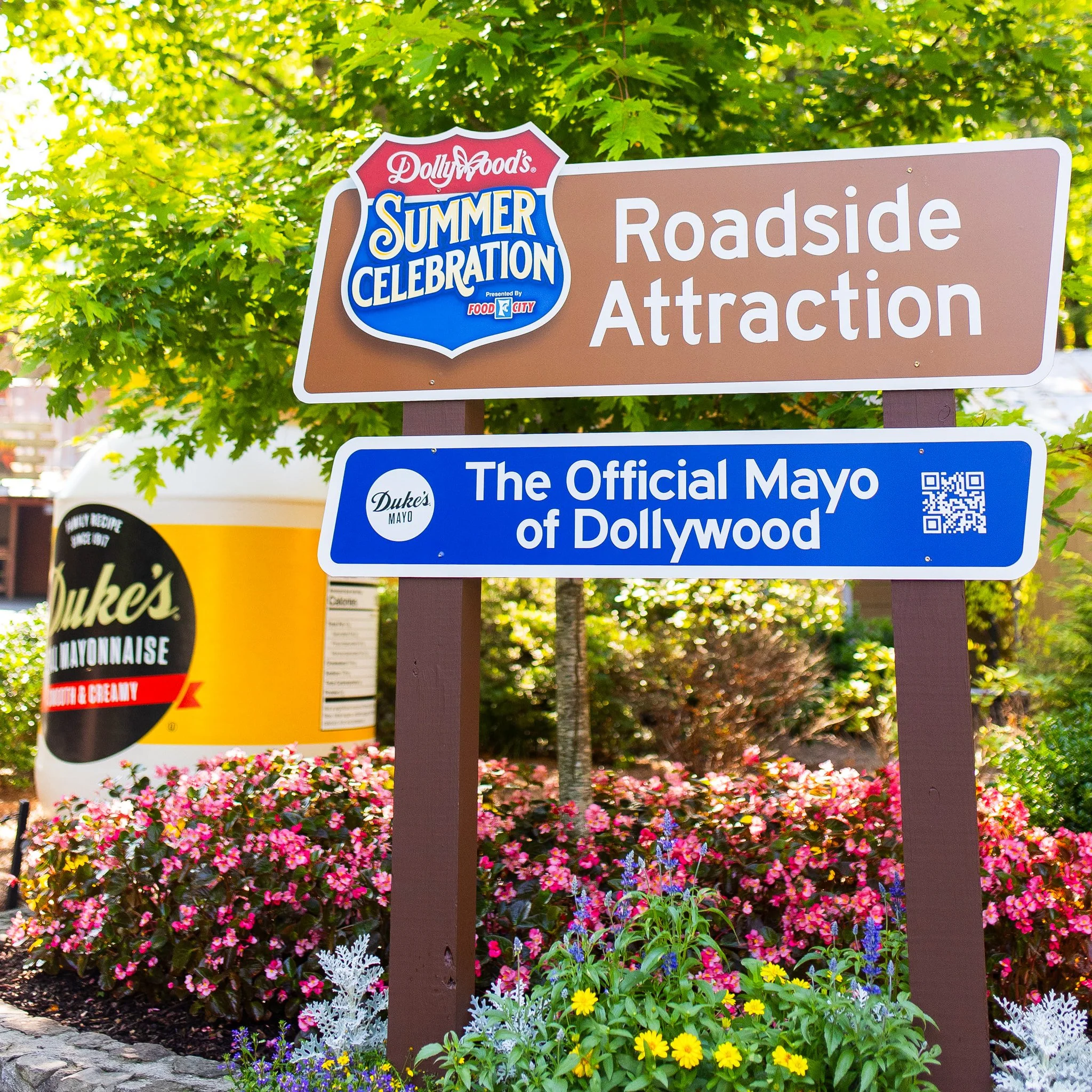 Sign promoting Dollywood's Summer Celebration roadside attraction, featuring official Mayo of Dollywood, with colorful flowers and a giant container of Duke's mayonnaise in the background.
