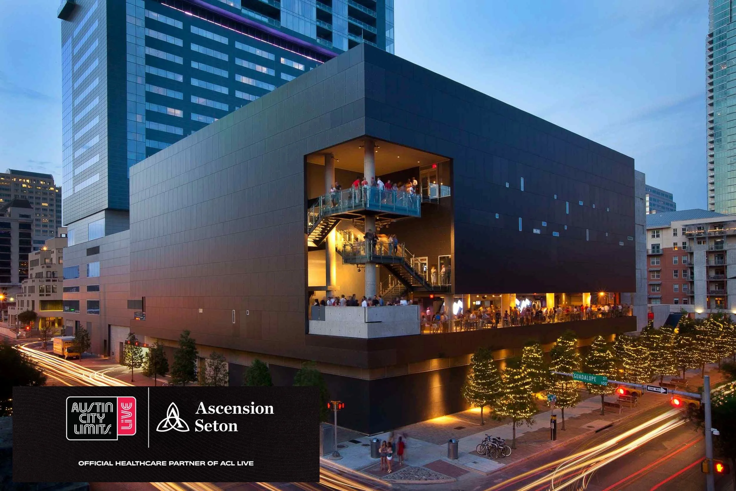 Modern multi-story building at dusk with illuminated outdoor balconies and a rooftop terrace filled with people. The lower level features a well-lit outdoor dining area with people and string lights on trees, and the street below shows light trails from moving vehicles.