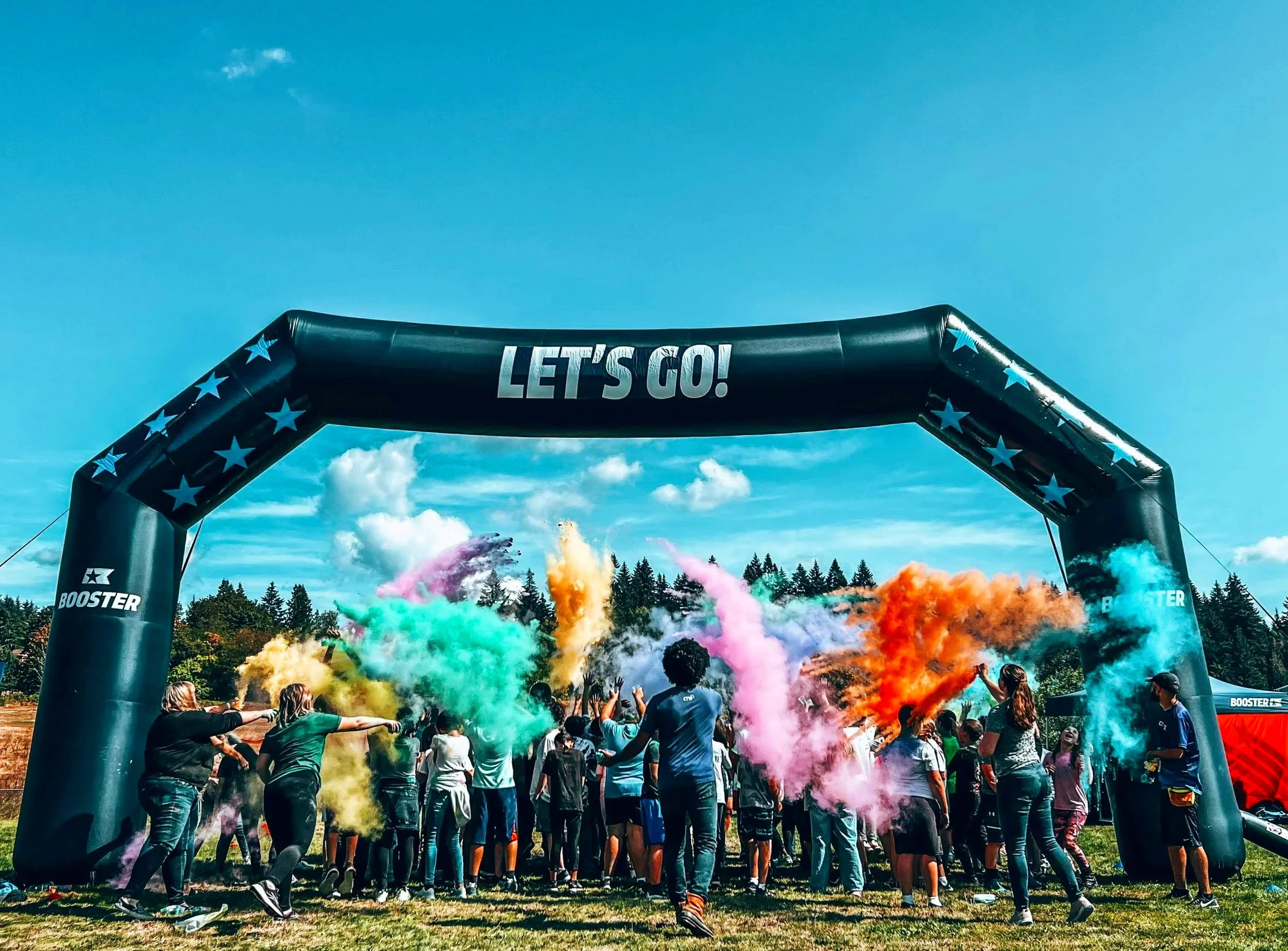 People participating in a color run event under a large inflatable arch that says 'Let's Go!', with colorful powder clouds in the air and a bright blue sky.