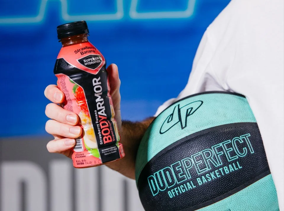 A person holding a BodyArmor strawberry banana flavored sports drink container, with an aqua and black Dude Perfect basketball sleeve on their arm.
