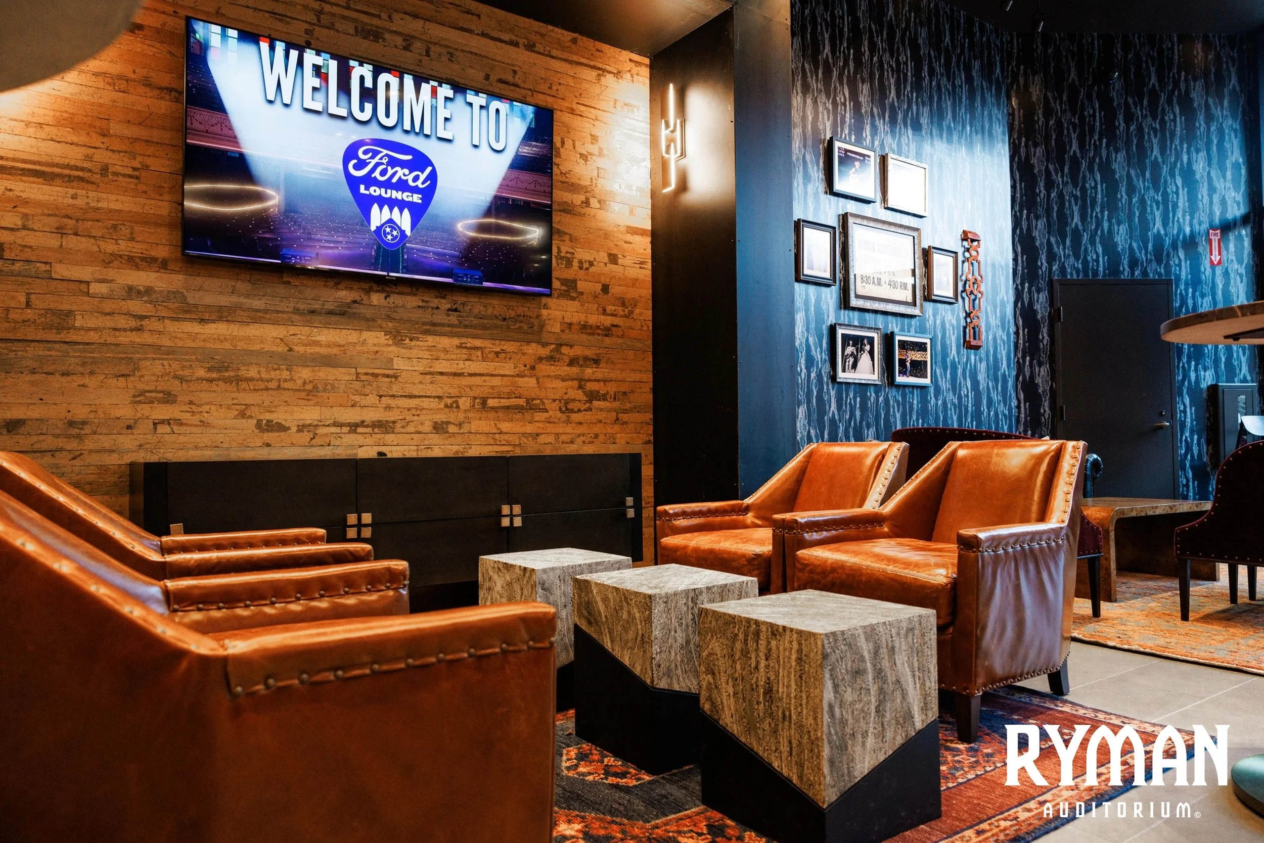A cozy lounge area with brown leather armchairs arranged around three marble-topped tables, a large flat-screen TV displaying a 'Welcome to Ford Lounge' message mounted on a brick wall, and framed pictures and artwork on the blue wall. The logo and the word 'RYMAN' are visible on the bottom right corner.