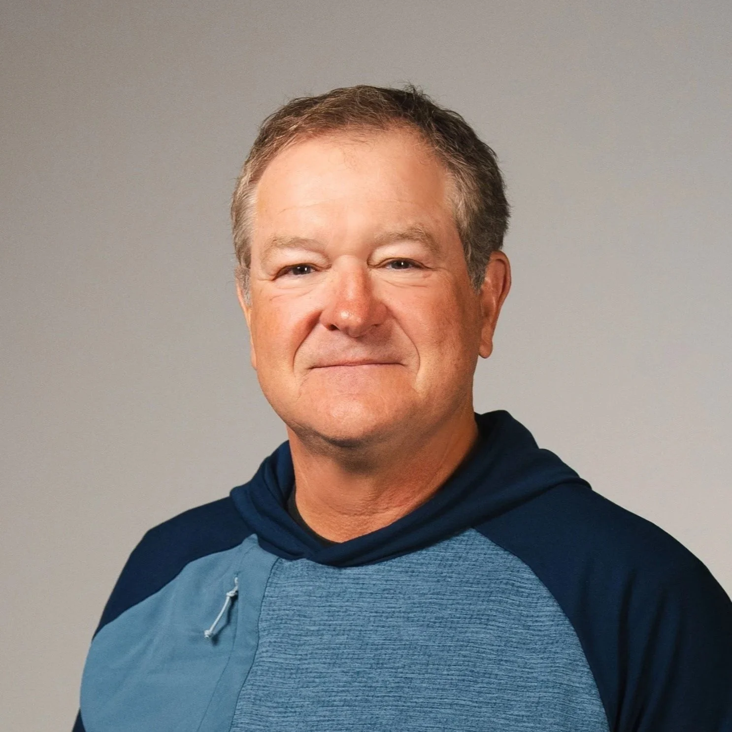 A middle-aged man with short hair, wearing a blue shirt, smiling softly, standing against a plain gray background.