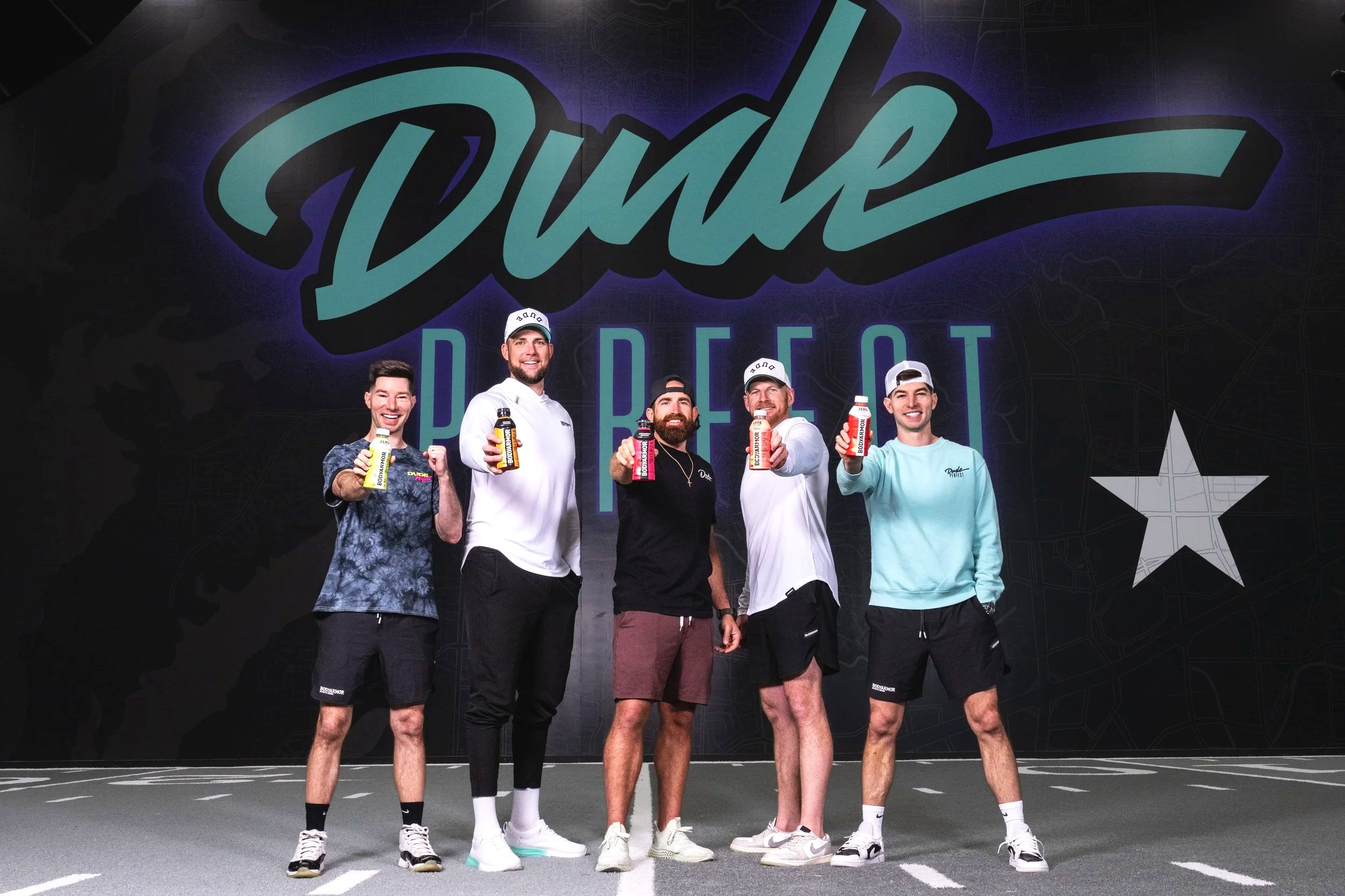 Five men standing in front of a background with the word 'Dude' and 'Dallas' written on it, holding bottles and smiling at the camera.