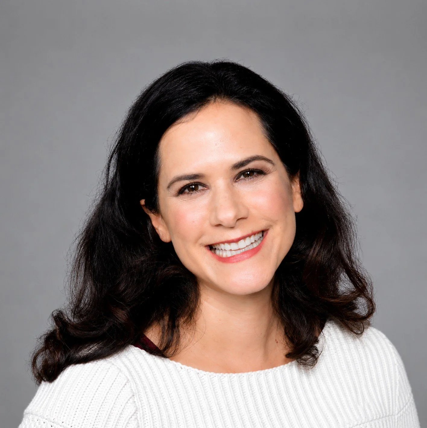Portrait of a woman with dark brown hair, smiling, wearing a red leather jacket and a white top, against a plain gray background.