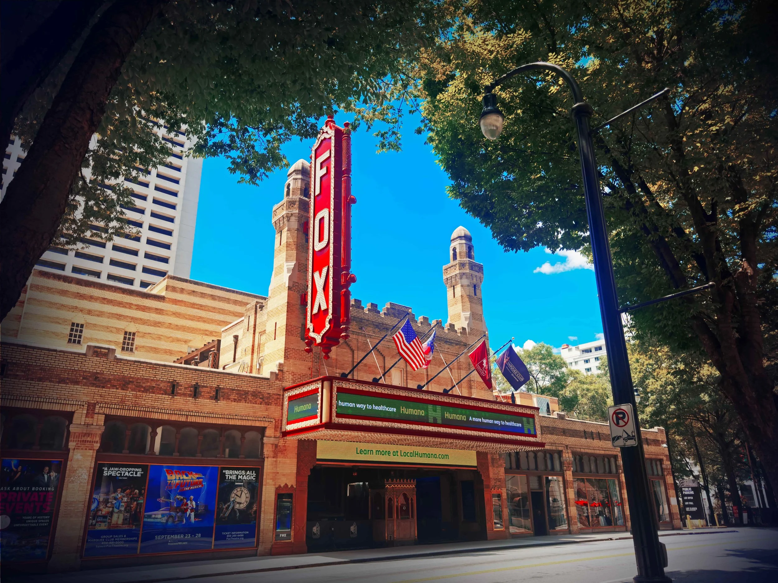 The Fox Theatre marquee sign on an old brick theatre building, with flags and posters on the sidewalk, and trees lining the street under a clear blue sky.