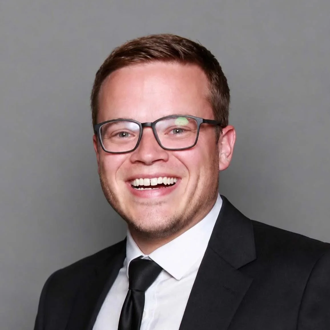 A smiling man with glasses wearing a gray zip-up jacket and a maroon collared shirt, posing against a plain gray background.
