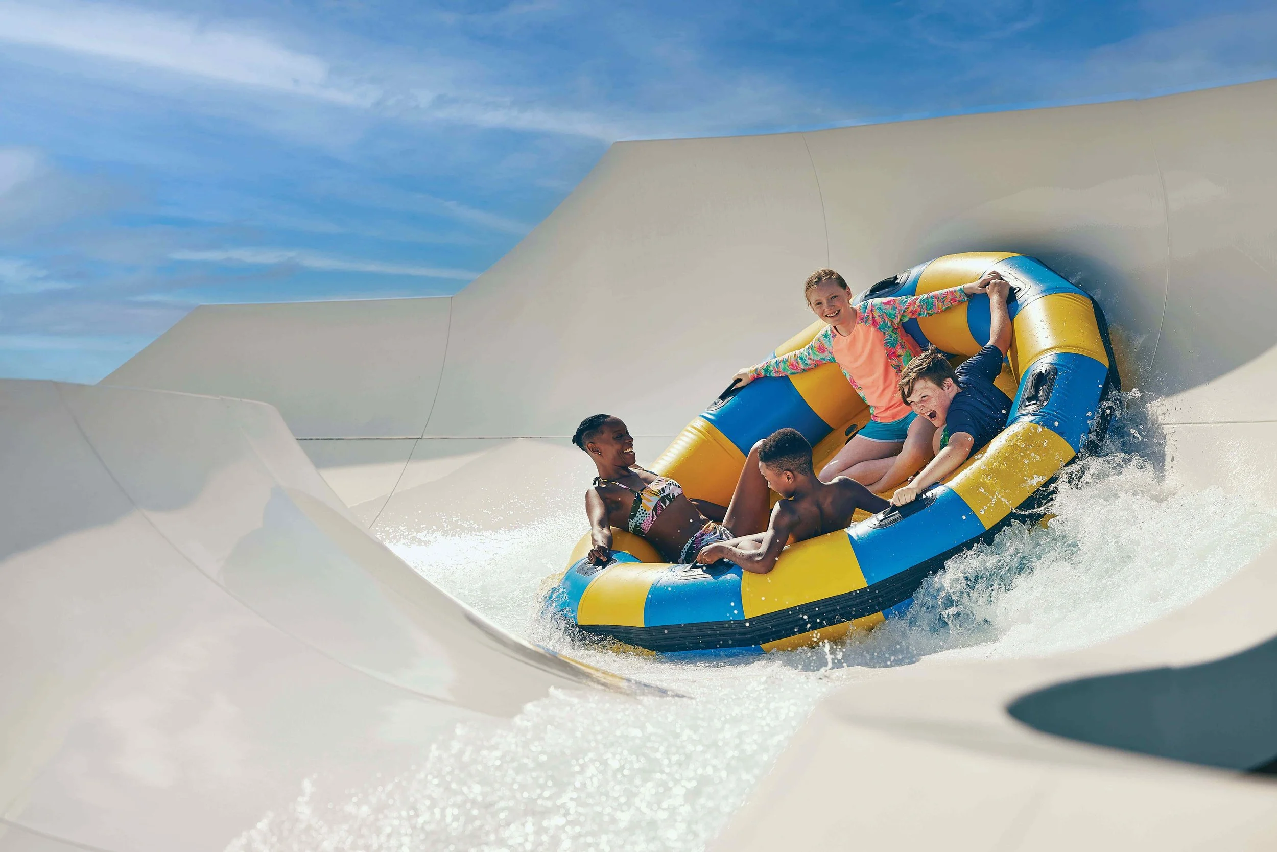 Children riding a yellow and blue inflatable raft on a water slide with a clear blue sky in the background.