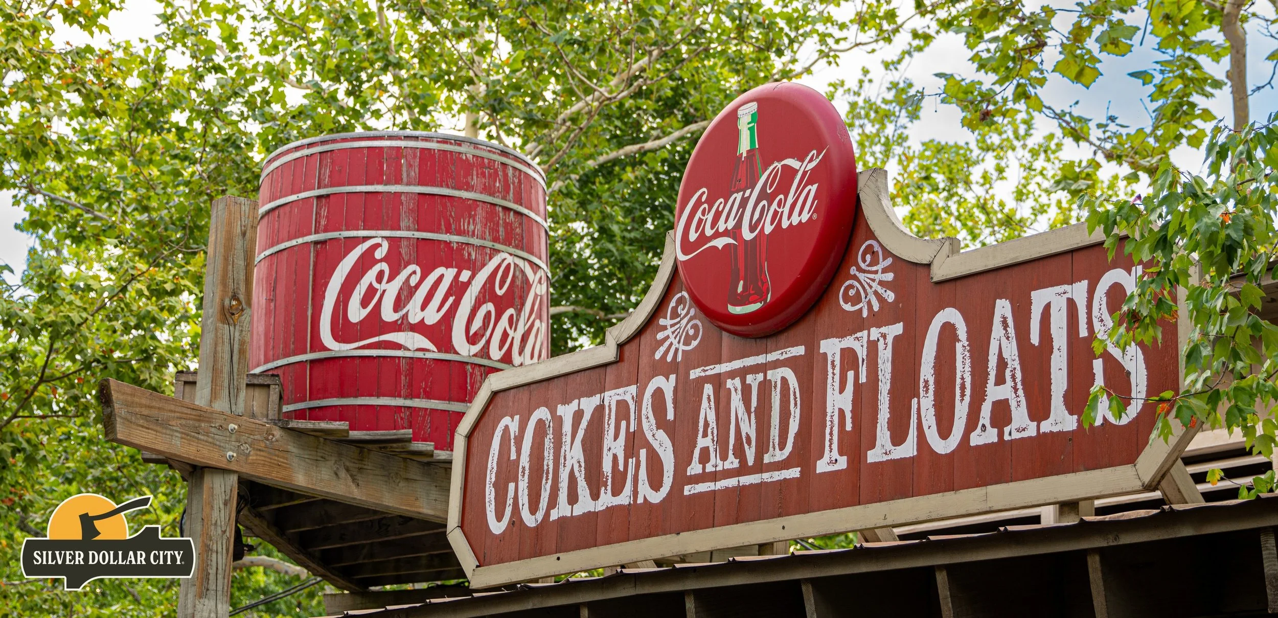 A vintage-style Coca-Cola sign with a red and white color scheme, featuring a round, illuminated Coca-Cola logo with a bottle image, and a rectangular sign reading 'Cokes and Floats' in white distressed lettering, surrounded by green trees.