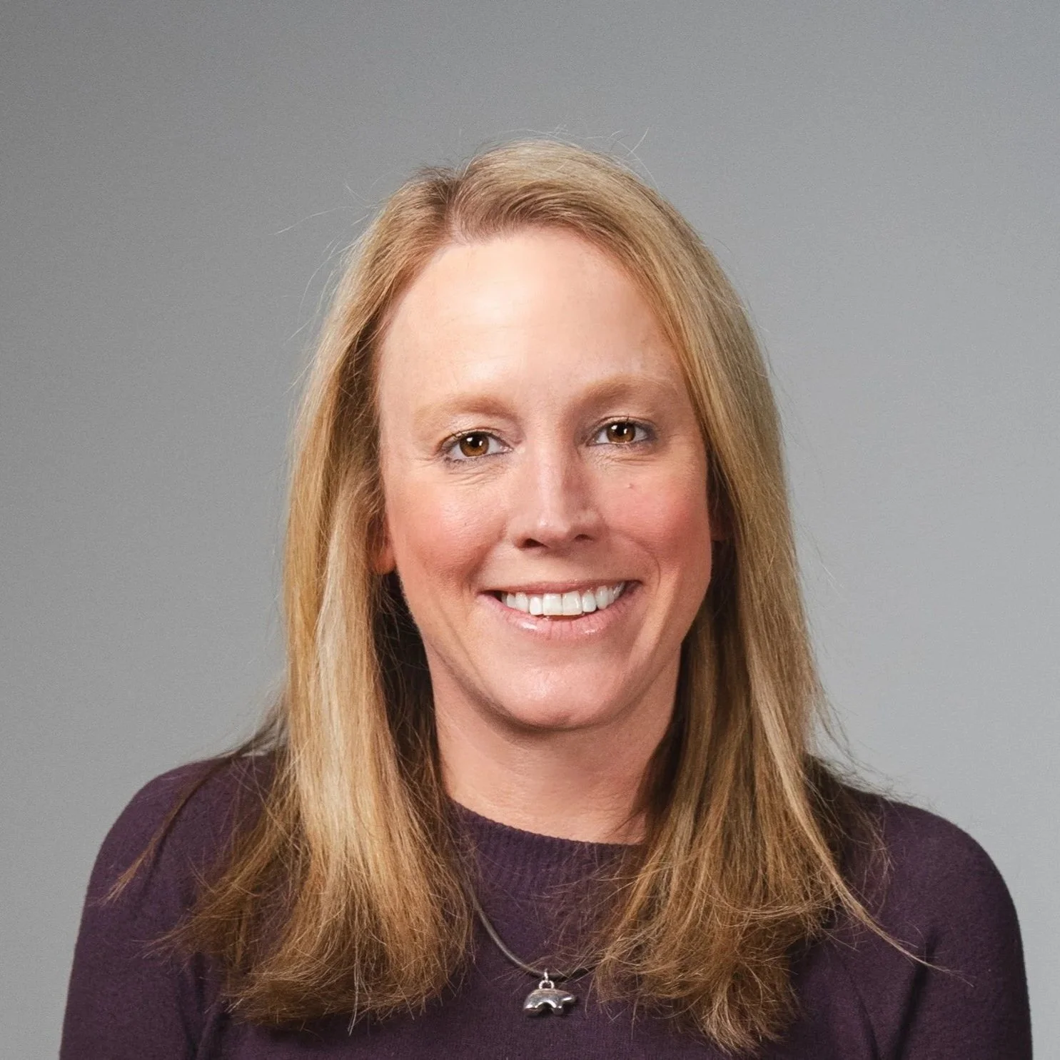 Headshot of a woman with long red hair, smiling, wearing a purple top and a necklace with a car pendant, against a plain gray background.