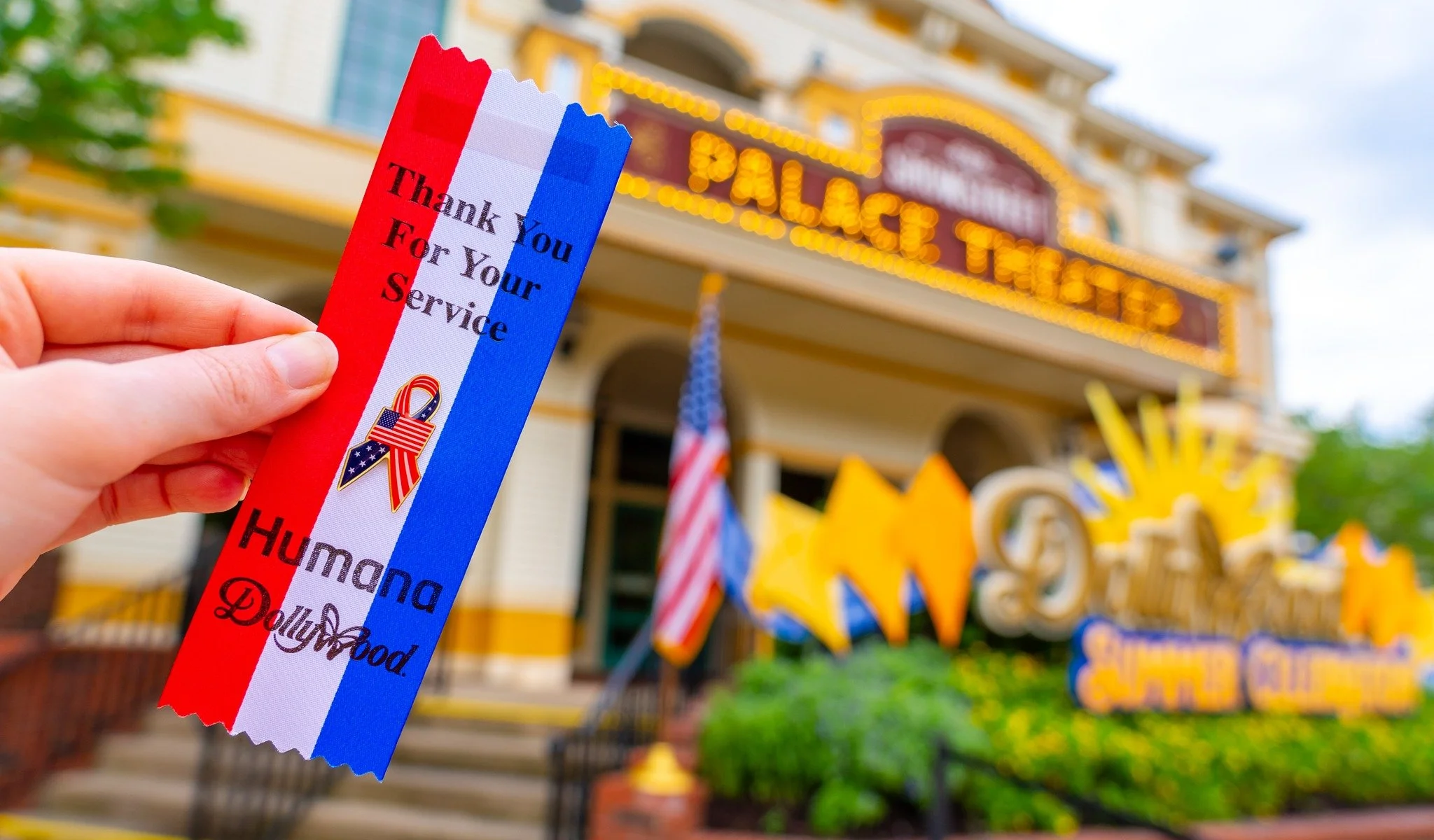 A person holding a ribbon with red, white, and blue colors and the words "Thank You For Your Service" and "HUMANA Dolly Parton" in front of a colorful storefront with signs and decorations.
