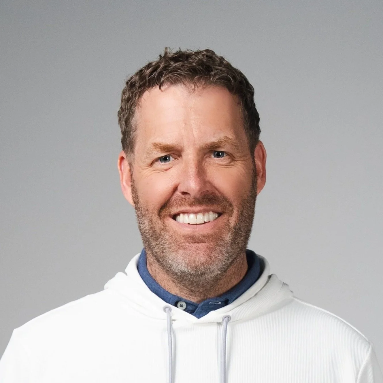 Smiling man with brown hair, beard, wearing a white hoodie and blue collared shirt against a plain grey background.