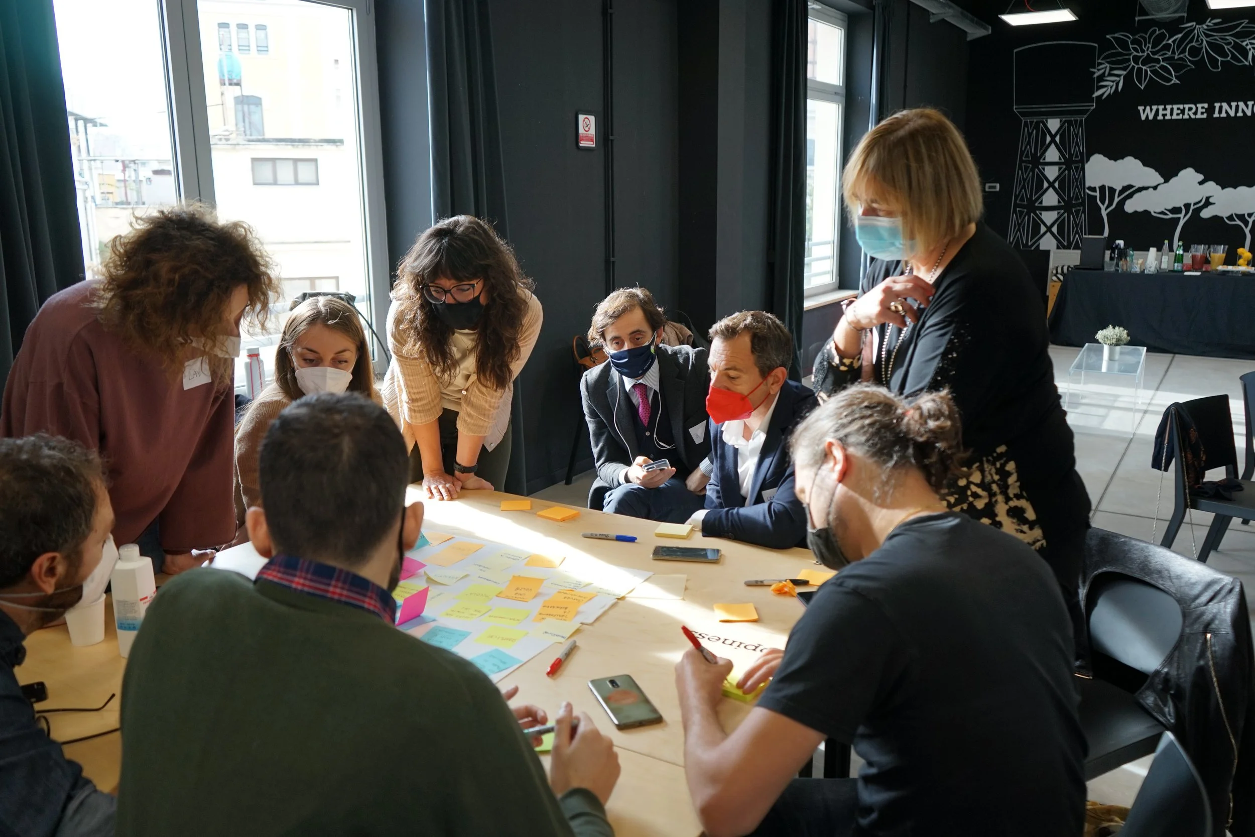Gruppo di persone che partecipano a un'attività collaborativa in una stanza moderna con grandi finestre e una parete decorativa con un diagramma. Tutti indossano mascherine e si concentrano su fogli e post-it sul tavolo.