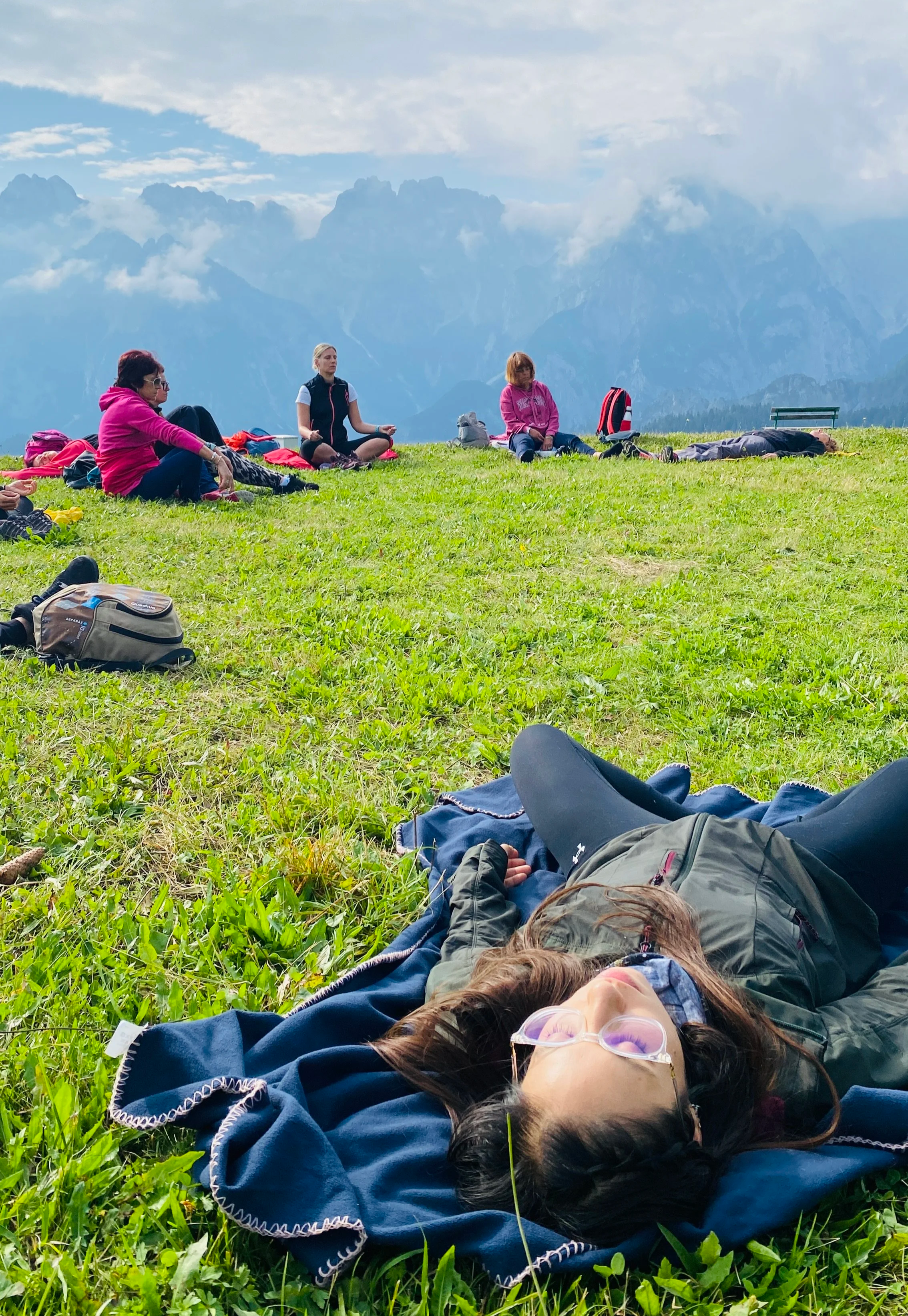 Gruppo di persone che praticano yoga o meditazione su un prato verde, con montagne sullo sfondo.