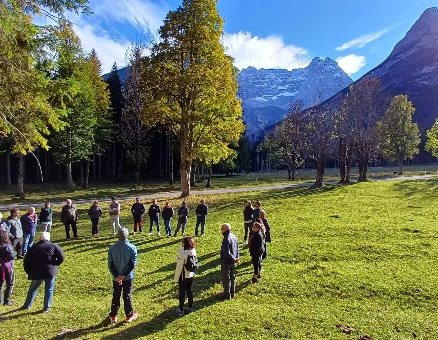 Gruppo di persone che si riuniscono in cerchio in un prato verde circondato da alberi e montagne con cielo sereno.