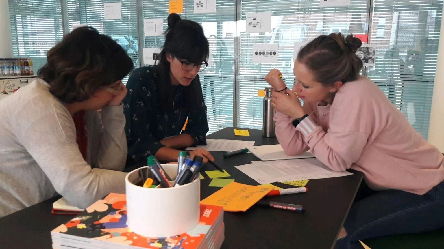 Tre donne sedute a un tavolo con materiale di cancelleria, mentre discutono o lavorano insieme in un ambiente di ufficio o studio.