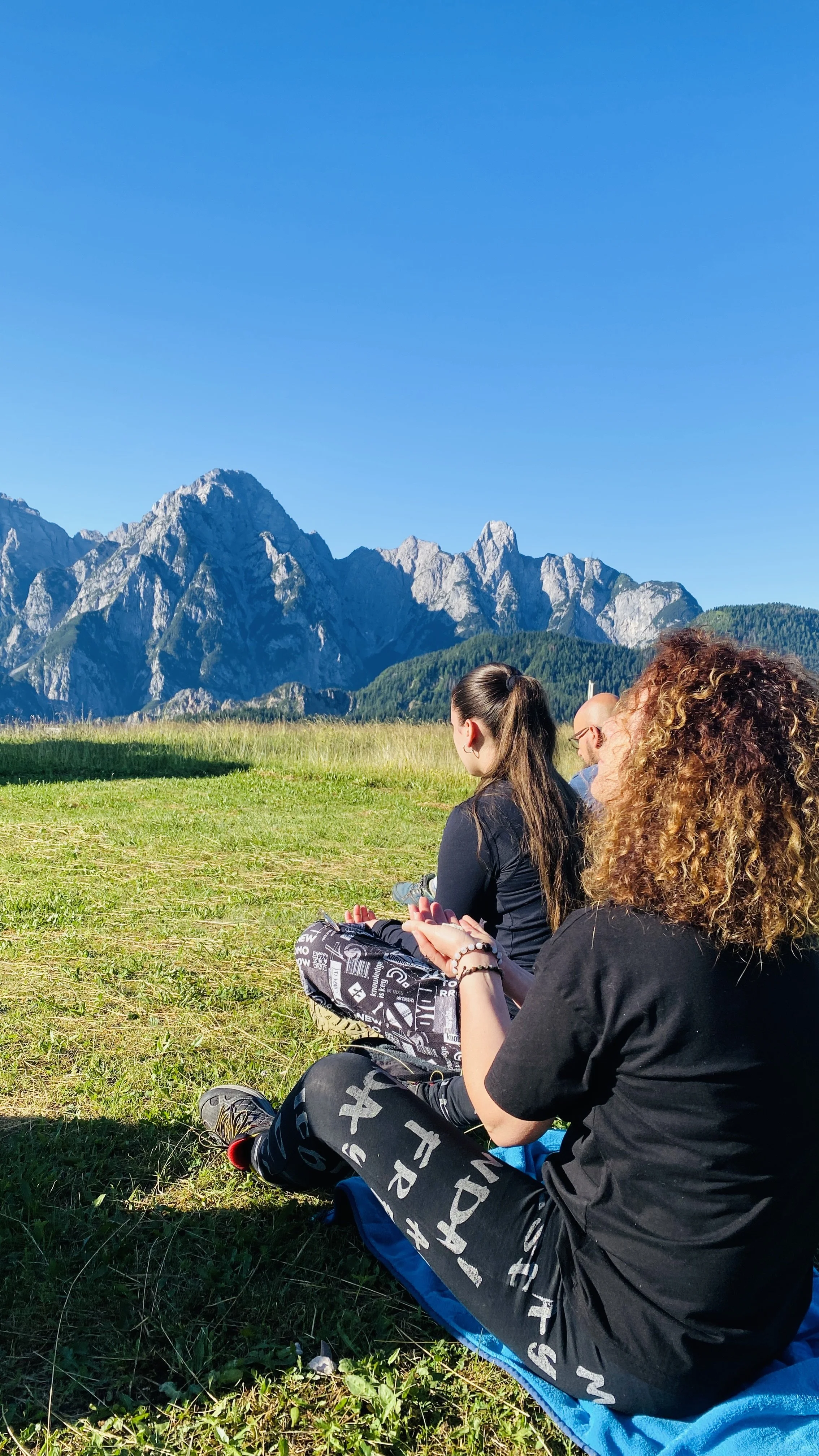 Tre persone sedute sull'erba che praticano meditazione con uno sfondo di montagne e cielo azzurro.