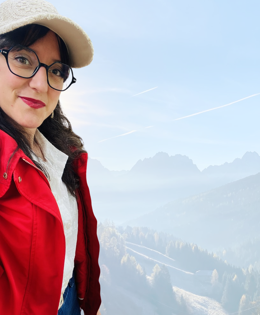 Donna con cappello, occhiali e giacca rossa davanti a un paesaggio di montagne e cielo blu.